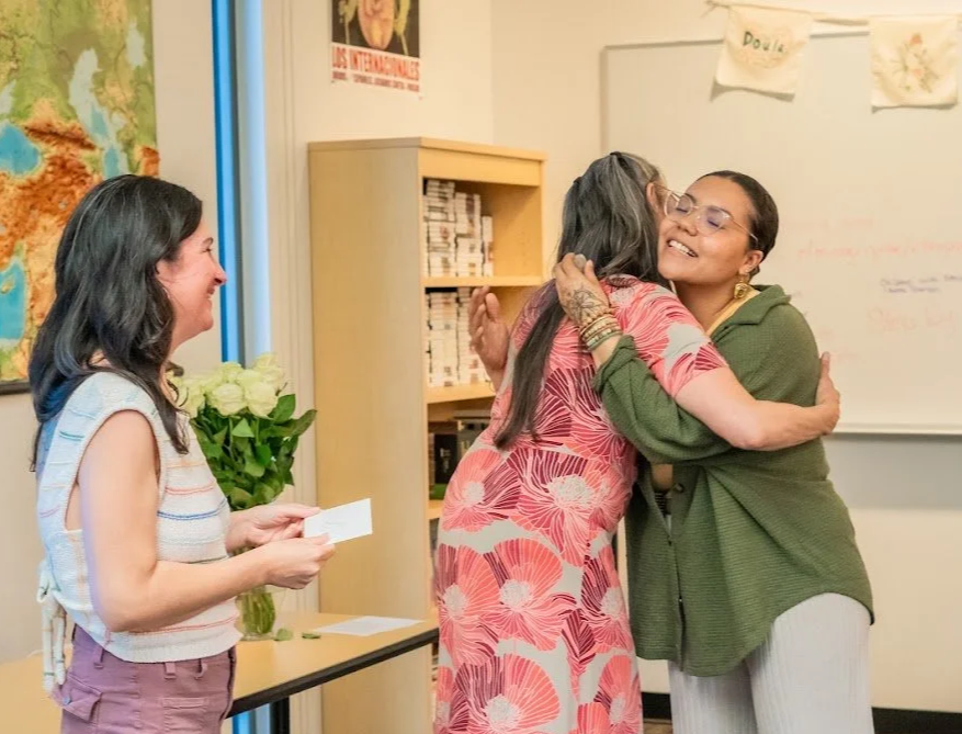 Three women in a classroom, two hugging and one smiling holding a card, shelves, a world map, and posters in the background.