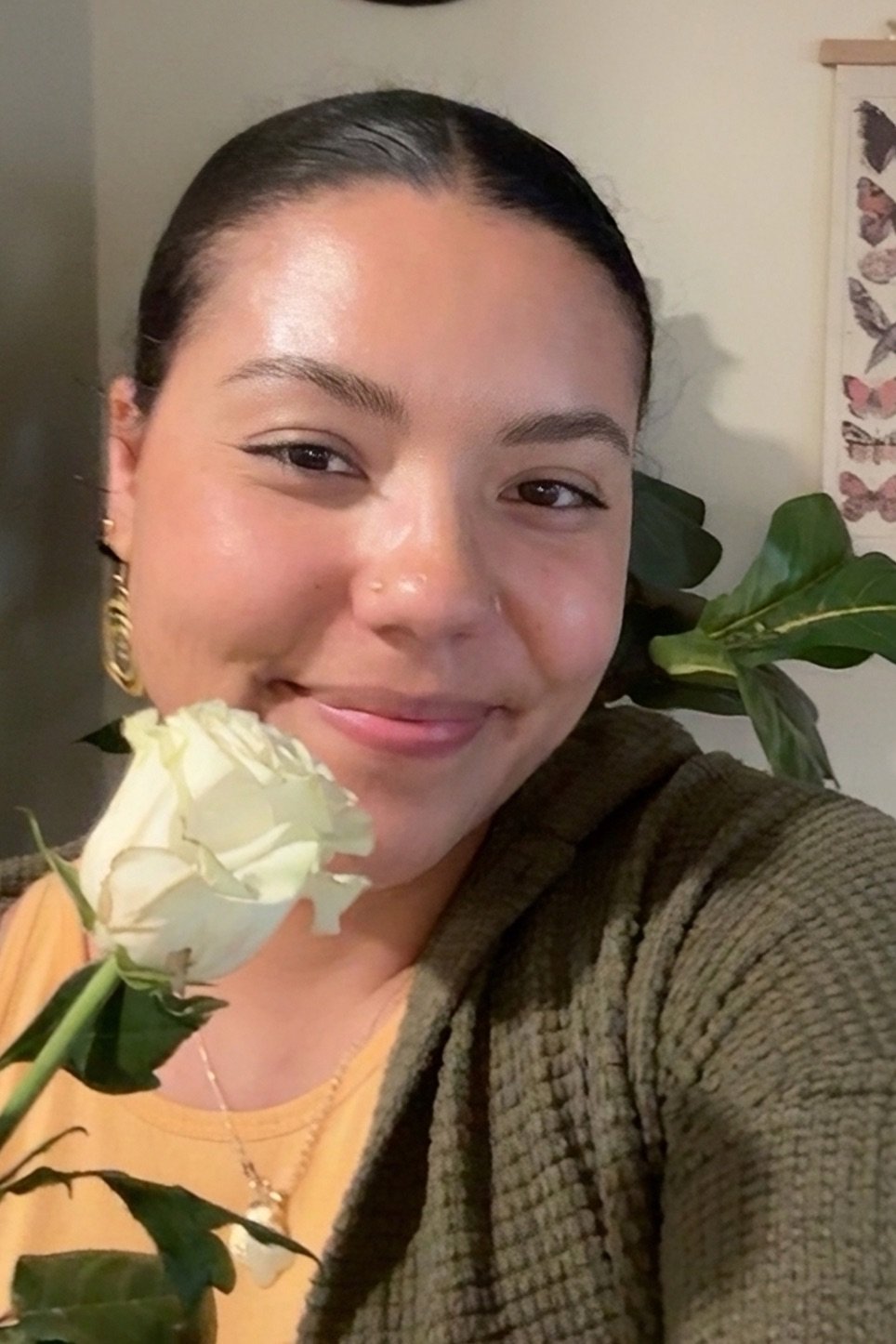 A woman smiling, holding a white flower near her face, with green plants and a butterfly poster in the background.