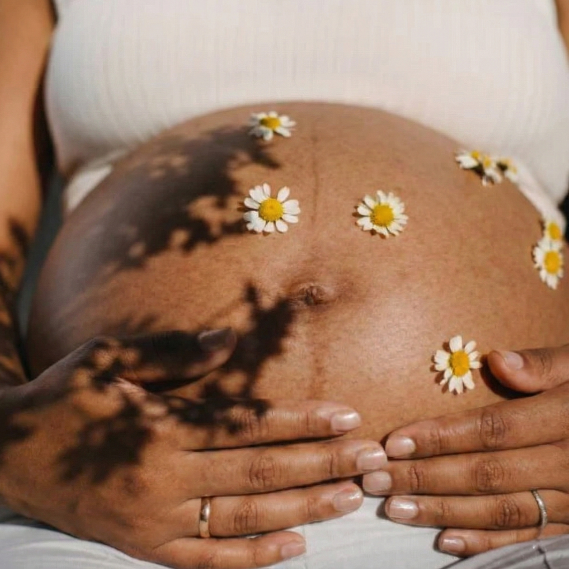 A pregnant belly with daisies on it, with shadows of flowers cast on the skin, and hands resting below.
