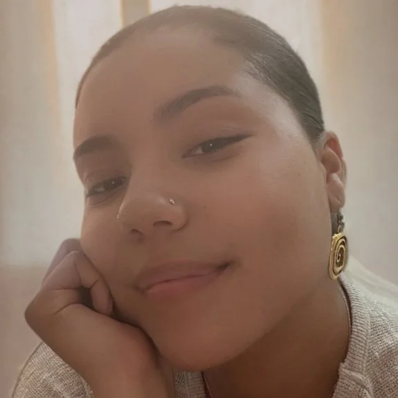 A close-up of a young woman with dark hair tied back, wearing a small nose piercing, gold spiral hoop earring, and a light-colored top, resting her chin on her hand and smiling slightly.