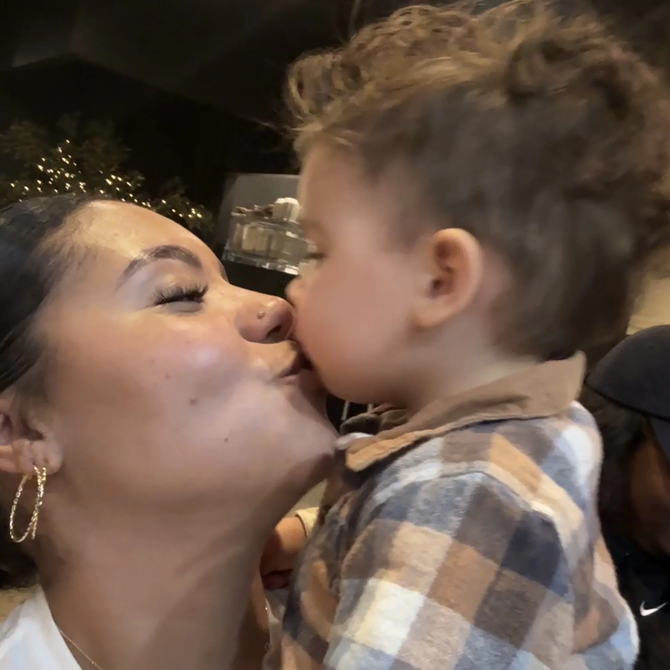 A woman and a young boy sharing a close moment, their faces touching, in a warm, indoor setting with a decorated Christmas tree in the background.