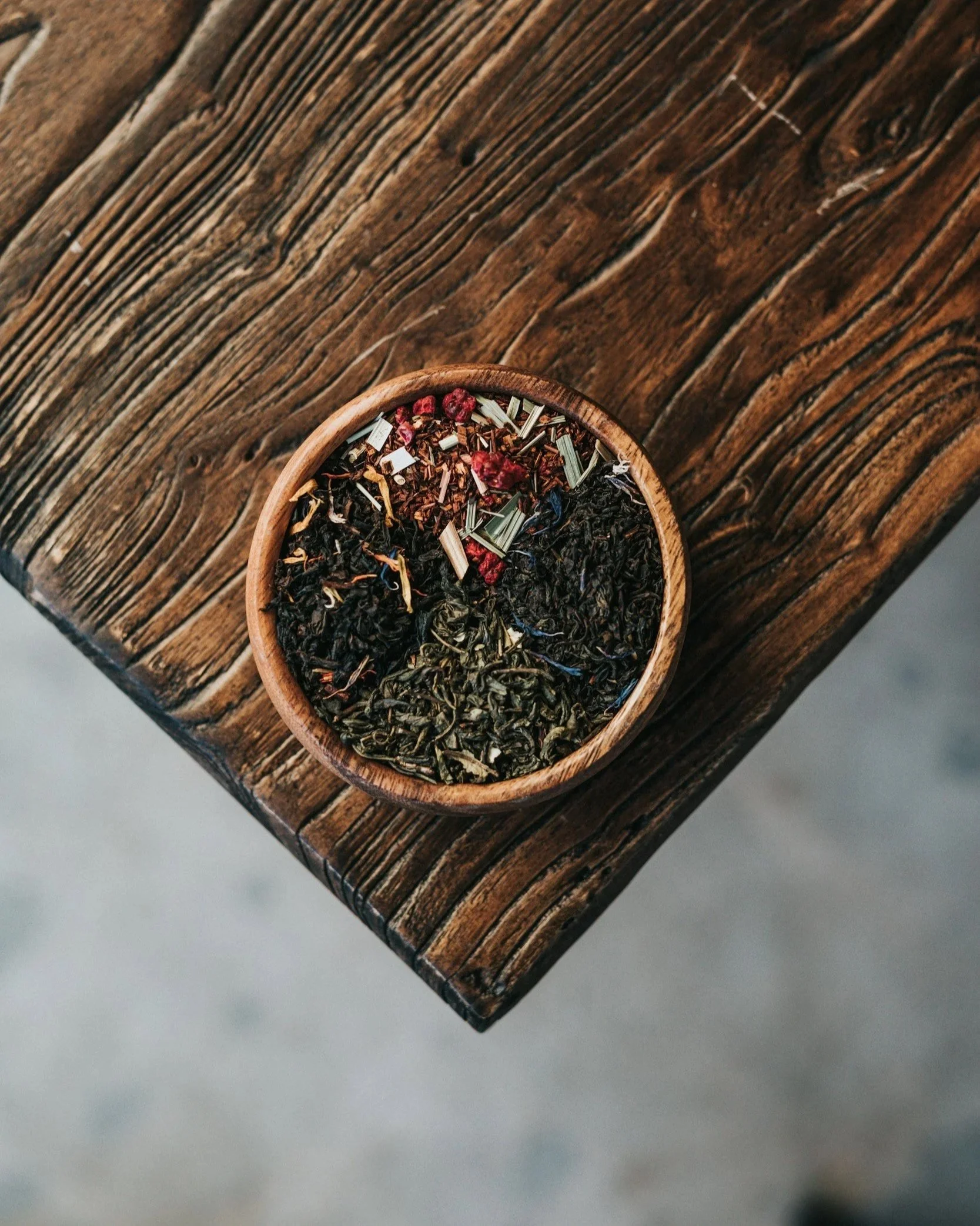 A round wooden bowl filled with loose leaf herbal tea, containing dried herbs and flower petals, placed on a rustic wooden surface.