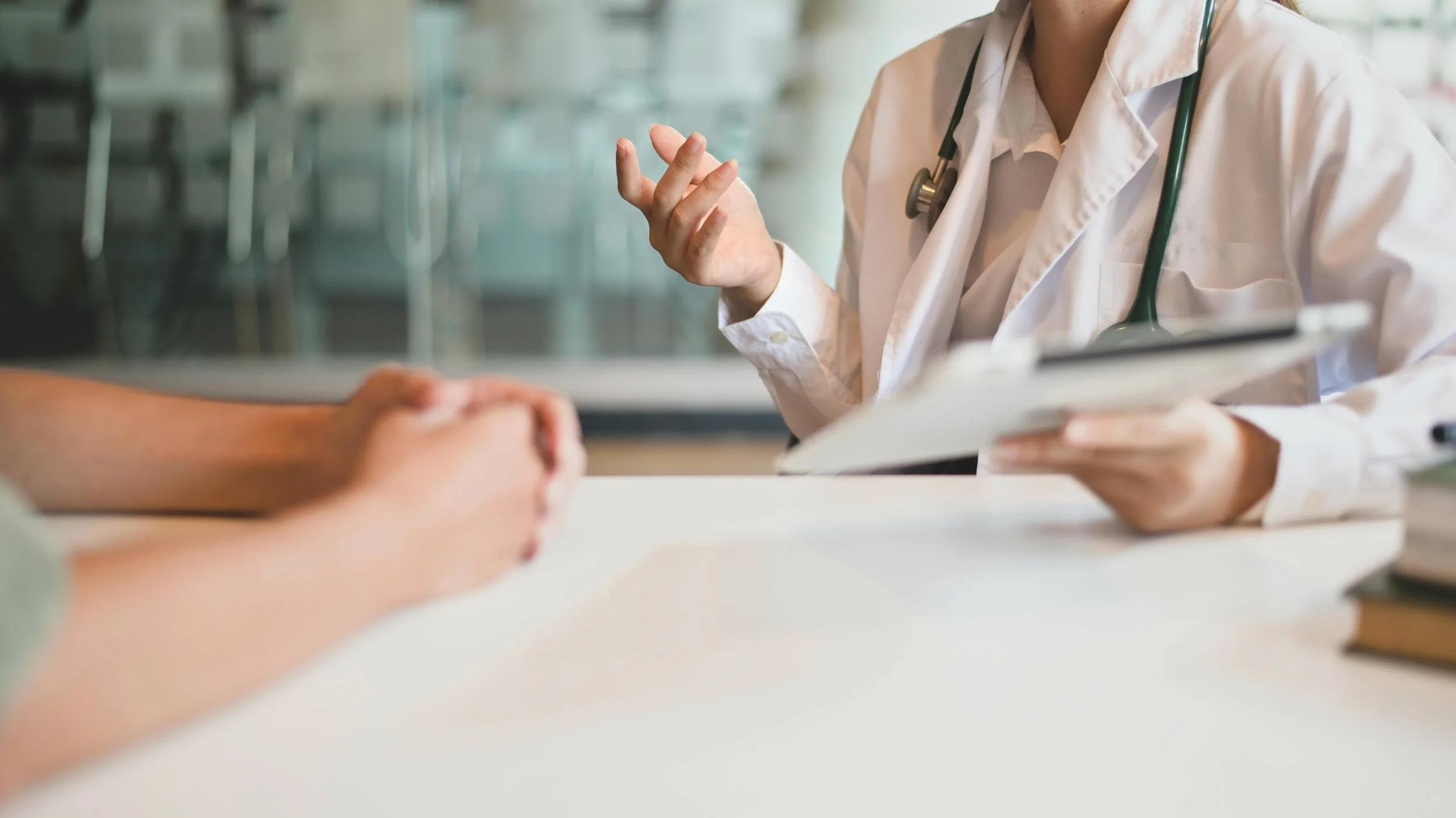 A doctor in a white coat and stethoscope talking to a patient, holding a clipboard, with the patient's hands resting on the table.