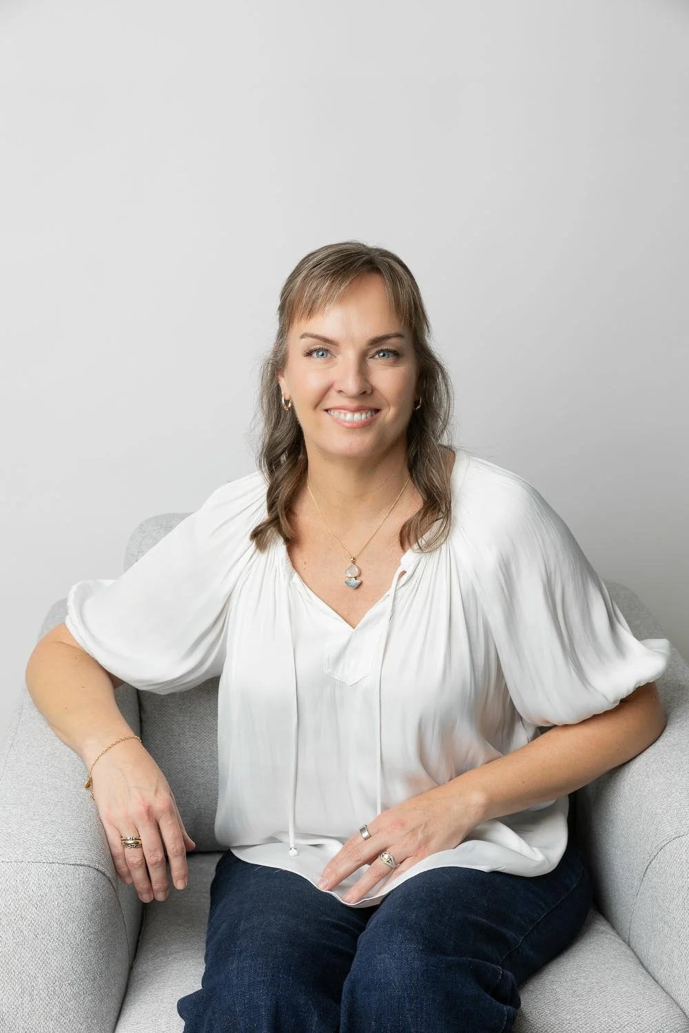 Portrait of Dr Ananta Kolesky sitting on a light gray sofa, smiling, wearing a white blouse, jeans, and jewellery, against a plain white background.