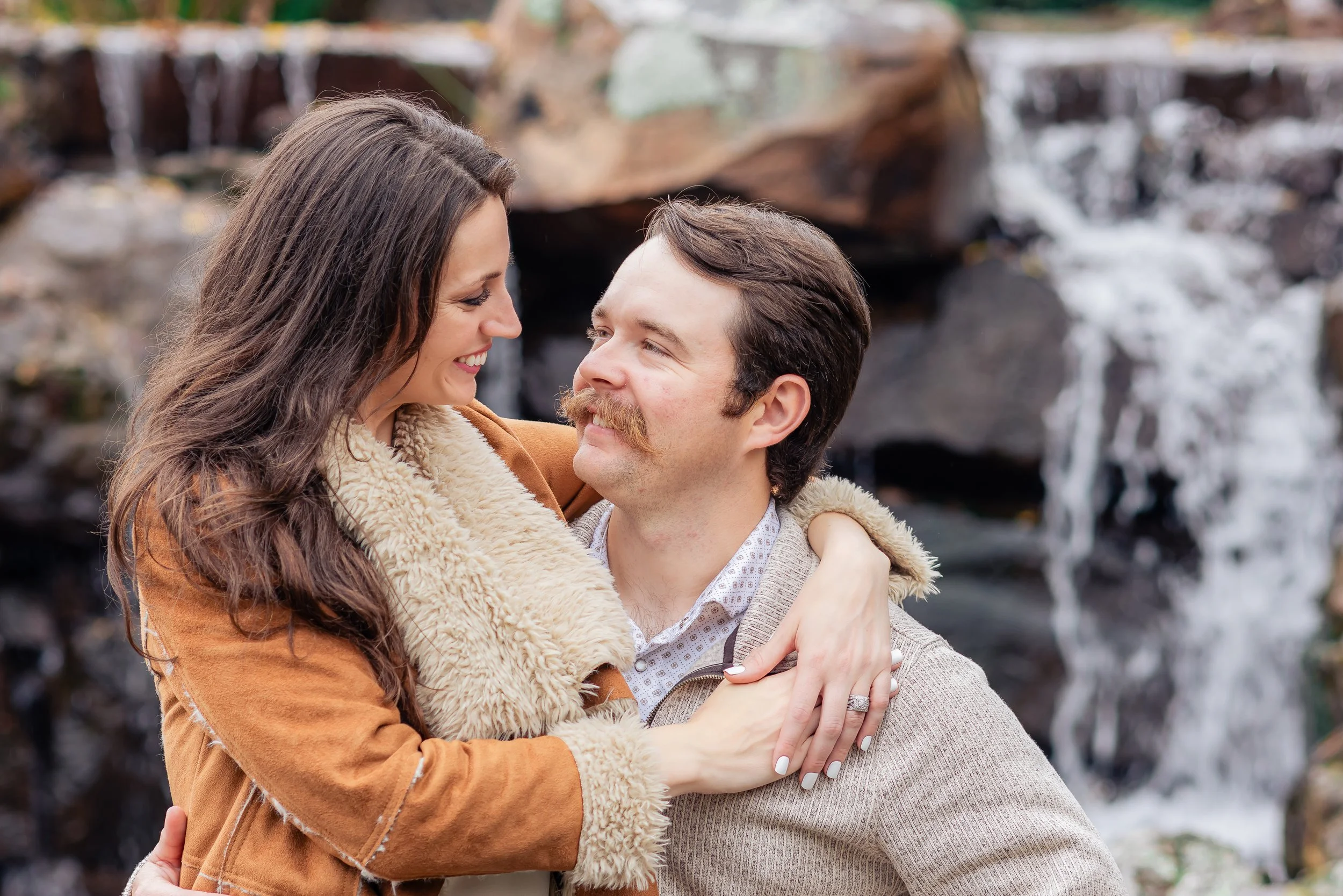 A couple embracing outdoors near a waterfall, smiling and looking into each other's eyes.