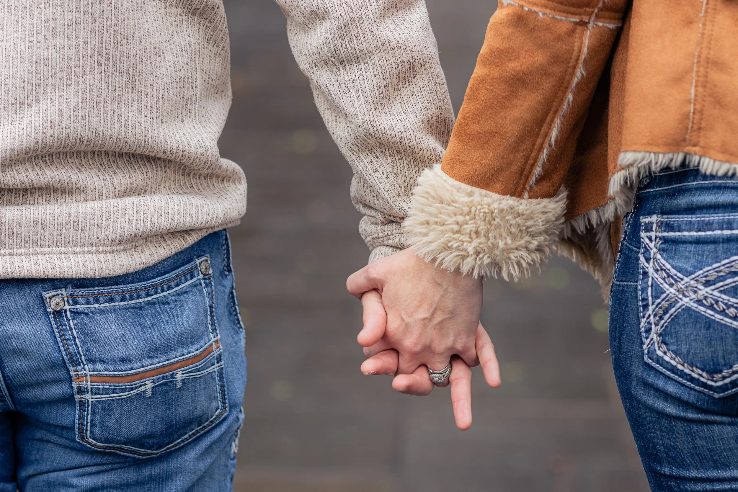 Two people holding hands, one wearing a beige sweater and the other wearing a tan jacket with fur trim, both in blue jeans.