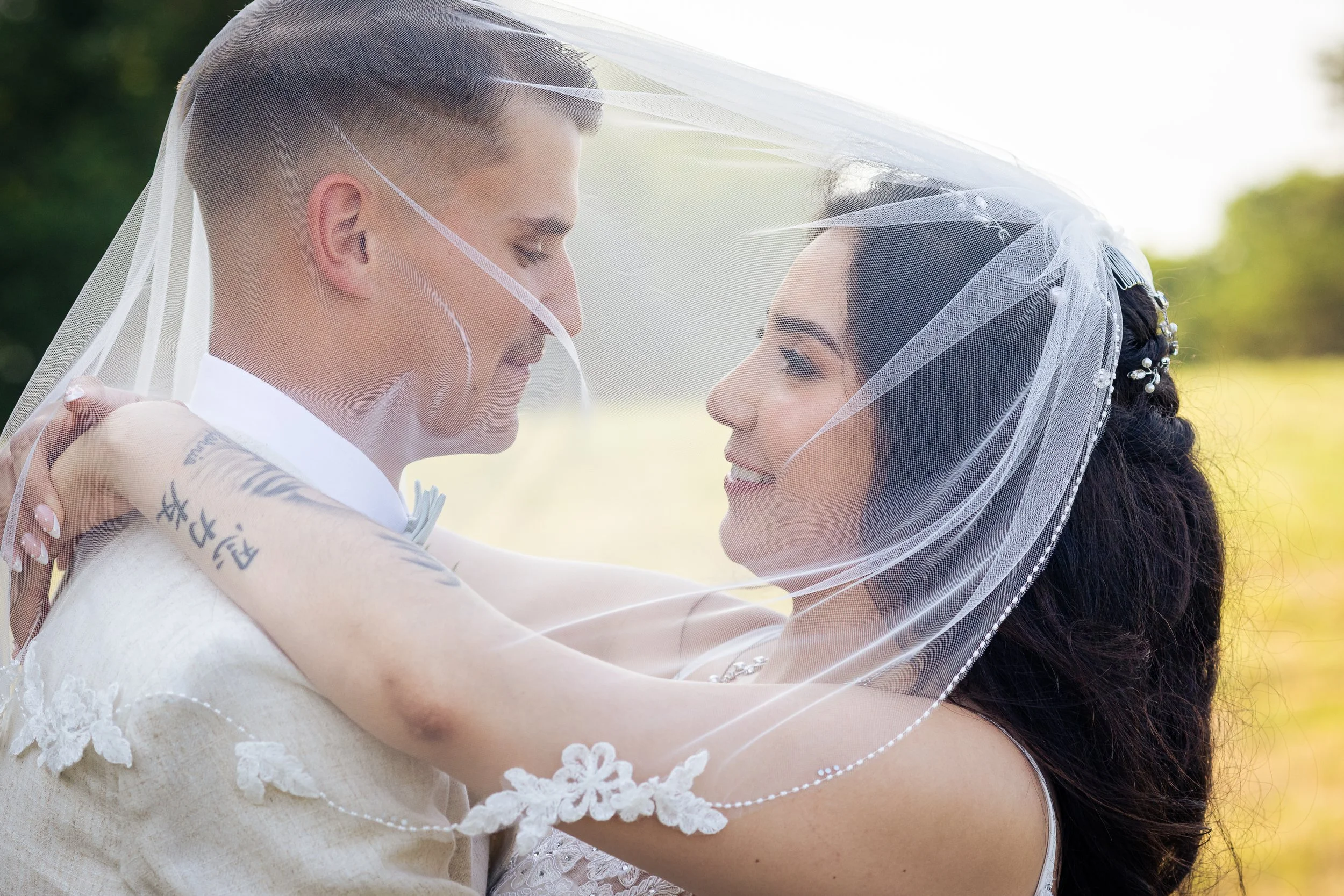 A bride and groom, face-to-face under a veil, smiling at each other outdoors on a sunny day.