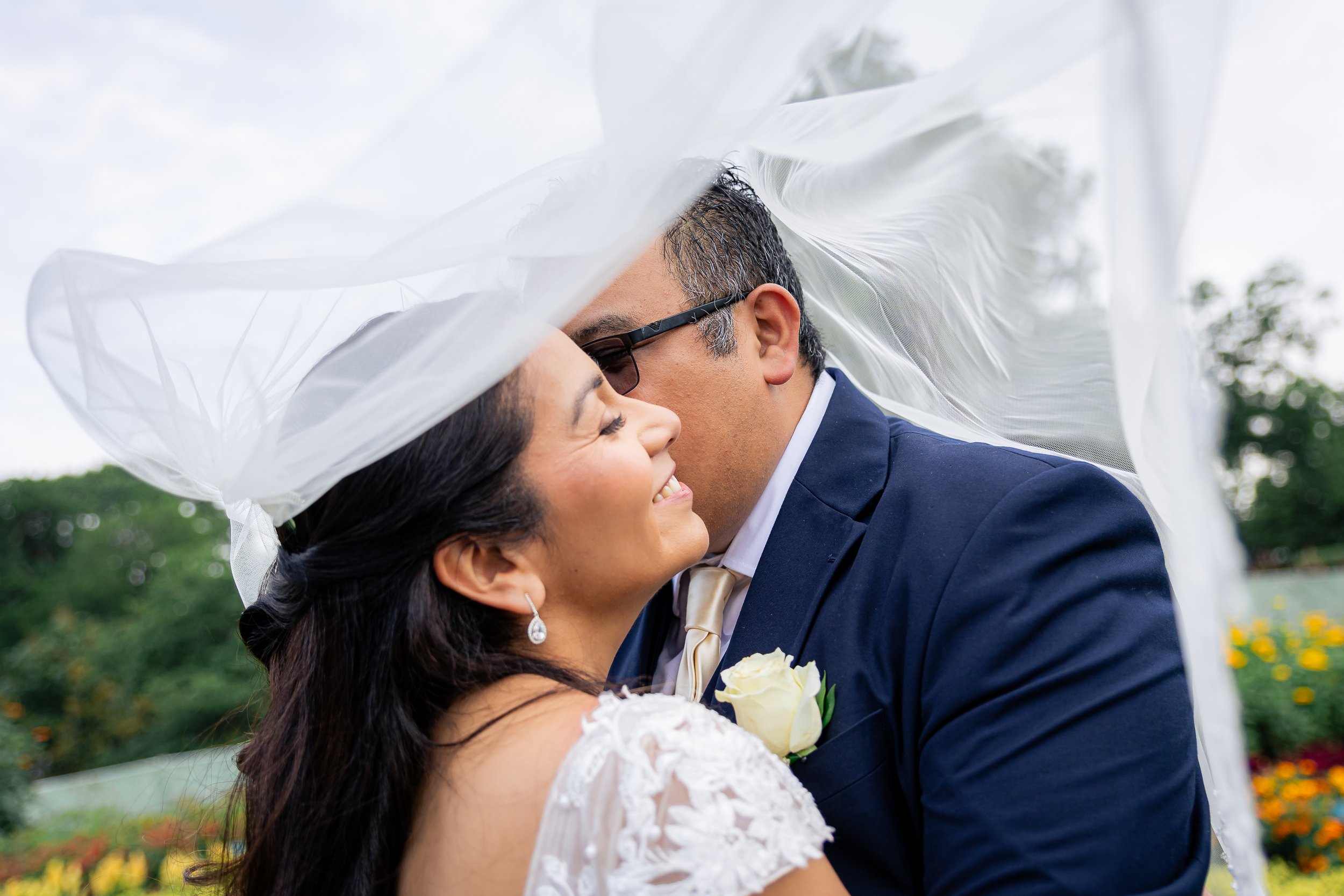 A happy couple, dressed in wedding attire, embracing outdoors under a white veil with green trees and flowers in the background.