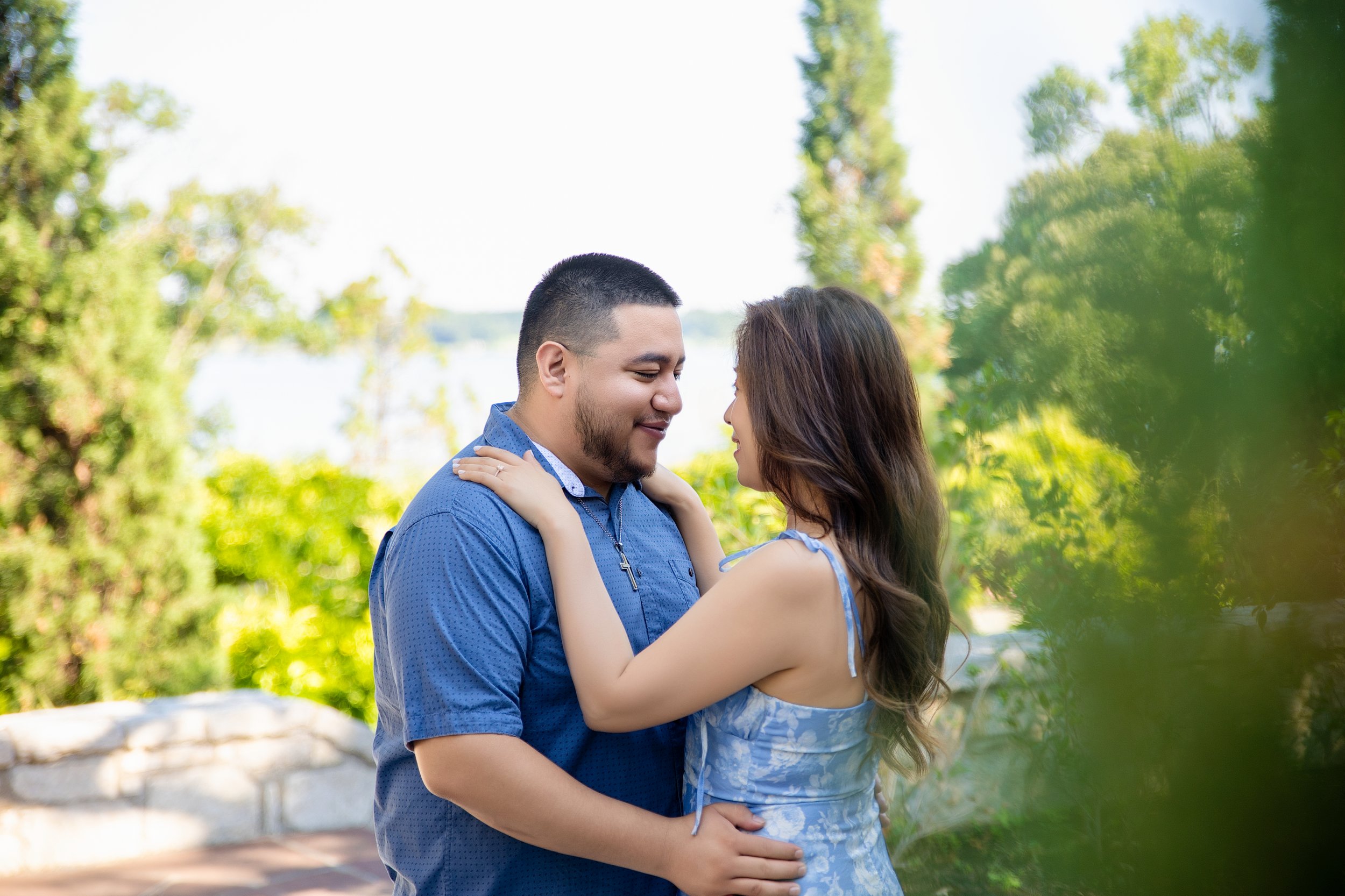 A couple holds each other closely outdoors on a sunny day, with trees and a lake in the background.