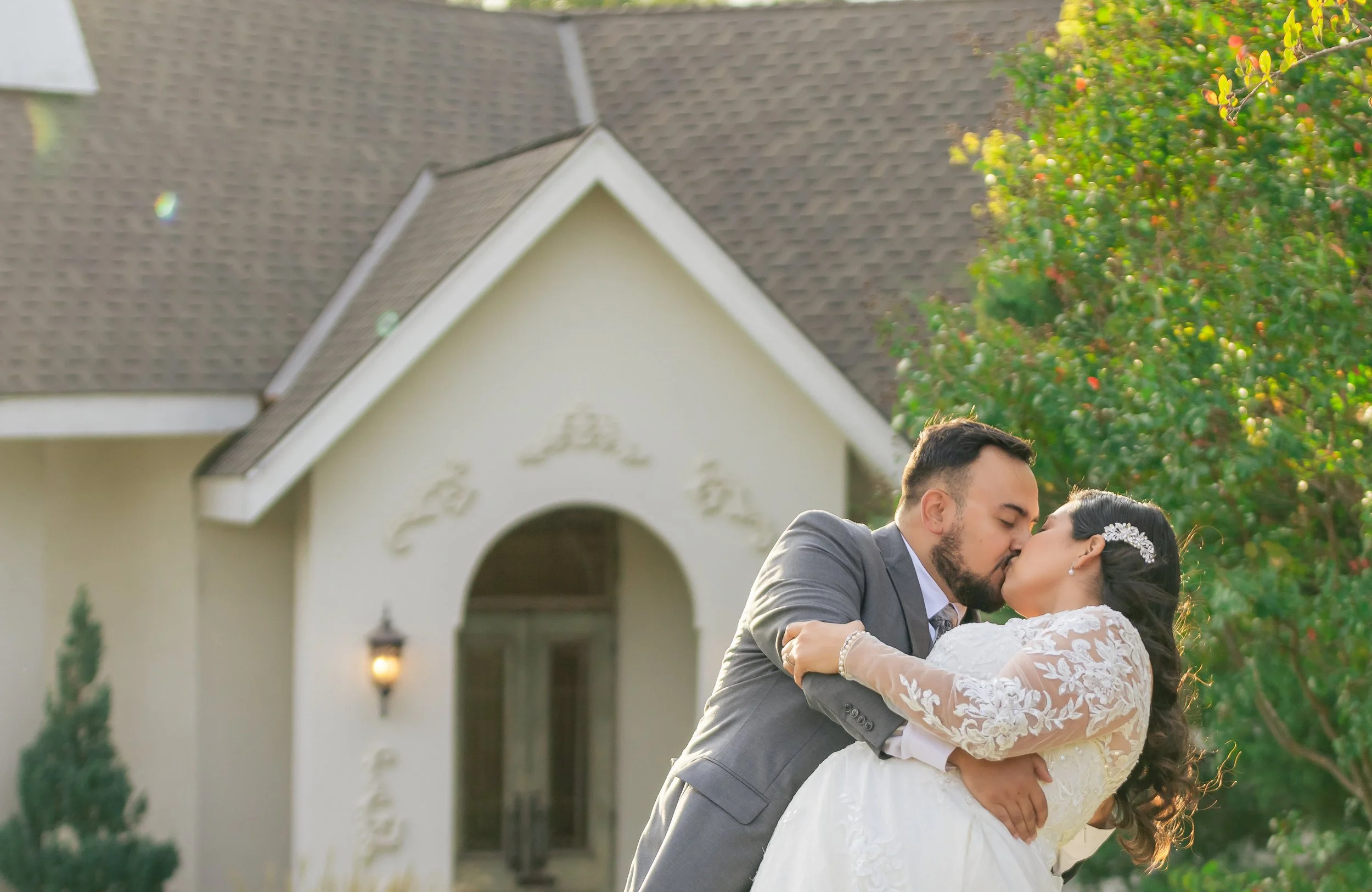 A newlywed couple sharing a kiss outdoors in front of a house with a peaked roof and arched entrance, surrounded by greenery.