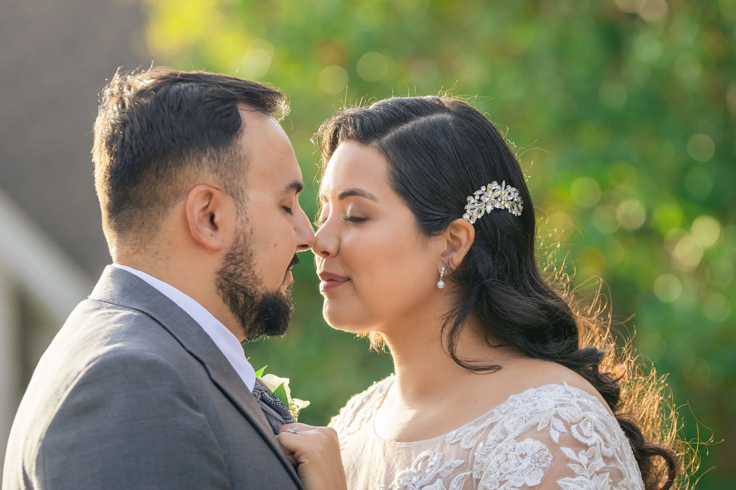 A bride and groom face each other closely outdoors, with their foreheads nearly touching, during a wedding ceremony.