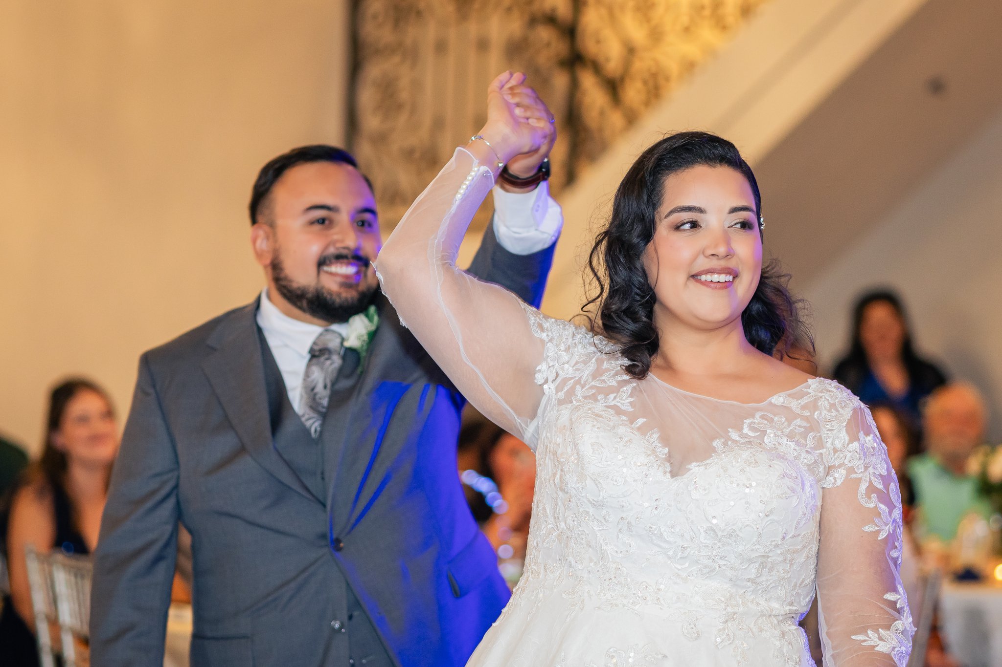 A bride and groom dancing at their wedding reception, with the groom holding the bride's hand and smiling, while the bride is smiling and looking ahead. Background guests are blurred.