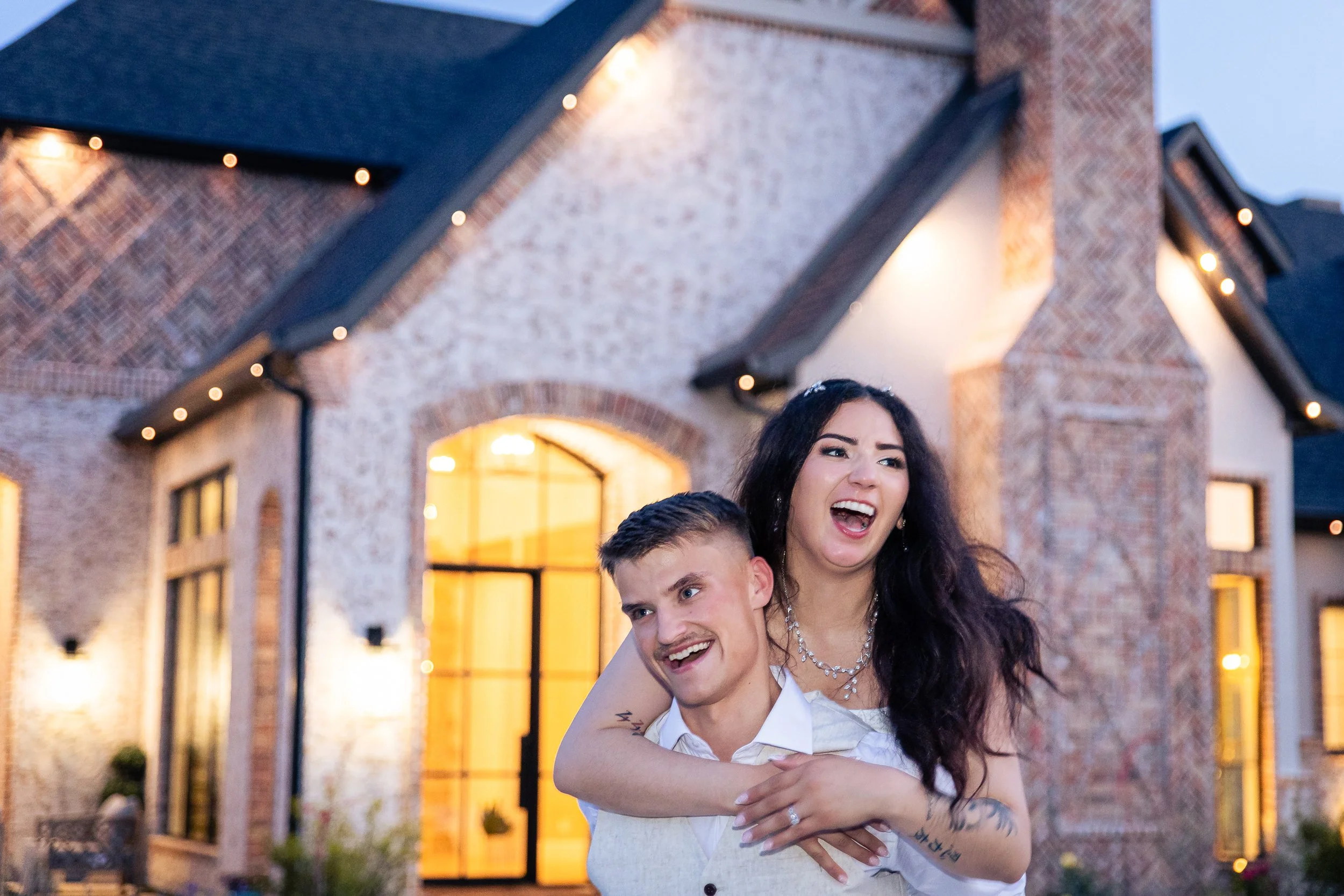 A woman and a young man are hugging and smiling in front of a brick house with arched windows and string lights hanging from the roof, during the evening.