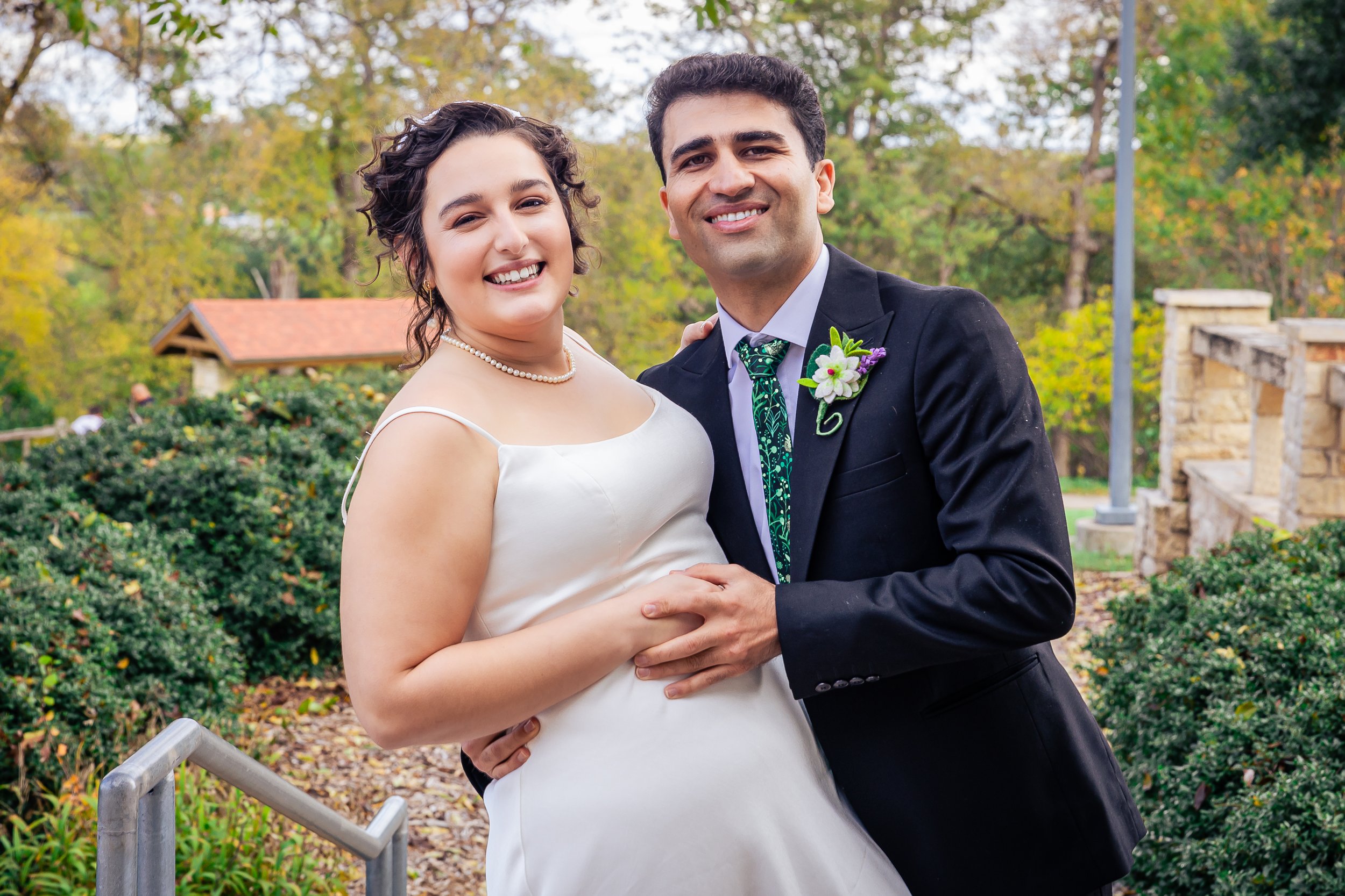 A smiling bride and groom posing together outdoors during a wedding, with trees and a stone structure in the background.