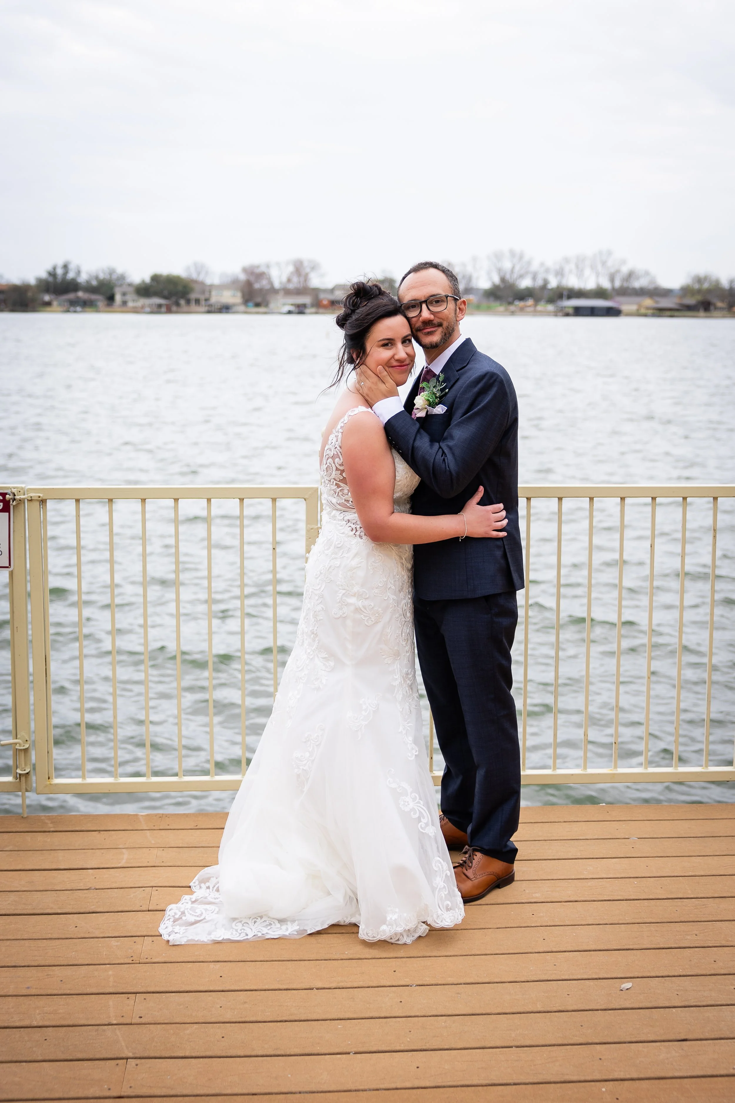 A newlywed couple stands on a wooden deck near a body of water. The bride is wearing a white lace wedding gown, and the groom is dressed in a navy suit. They are embracing with the bride smiling slightly and the groom smiling, with a cloudy sky above and houses visible across the water.
