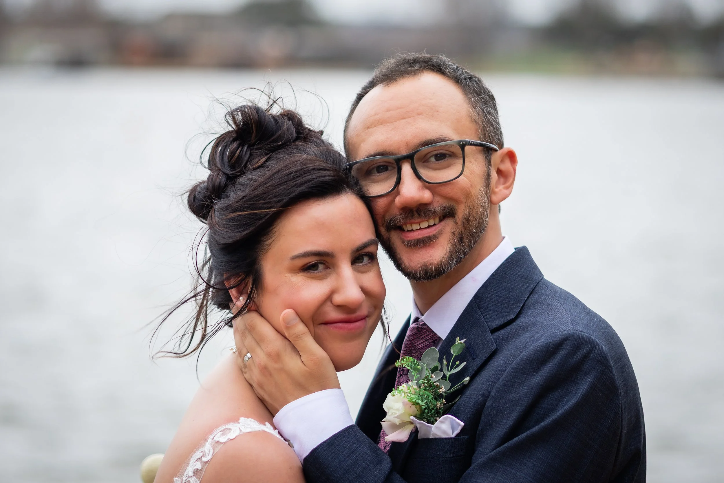 A close-up of a happy couple embracing outdoors near a body of water, dressed in wedding attire. The woman has dark hair styled in an updo, and the man has short dark hair, glasses, and a beard.