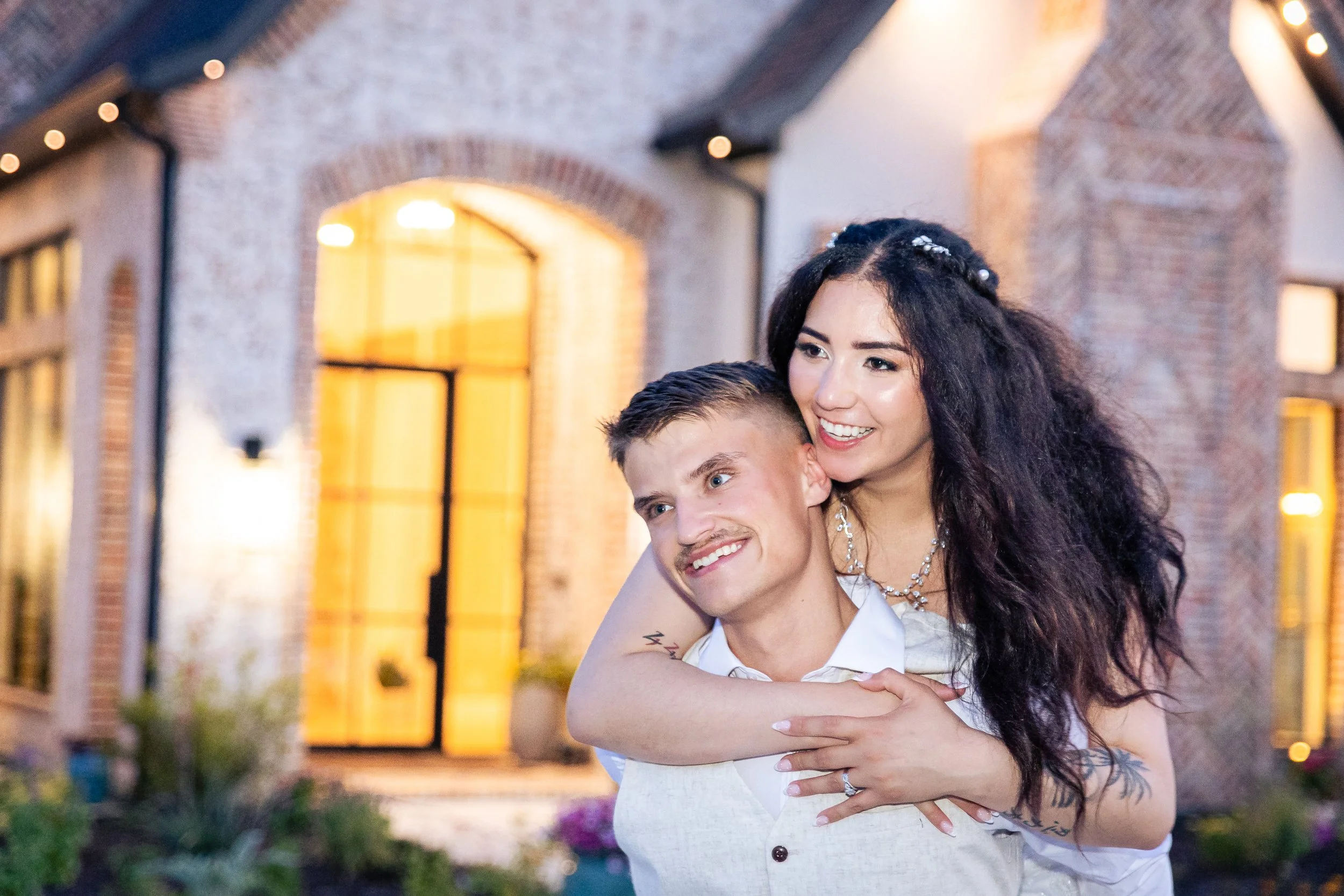 A smiling woman with long curly dark hair and a man with short light brown hair hugging in front of a brick building with arched windows at dusk. The woman is wearing jewelry and a white dress, the man a white shirt, and there are fairy lights in the background.
