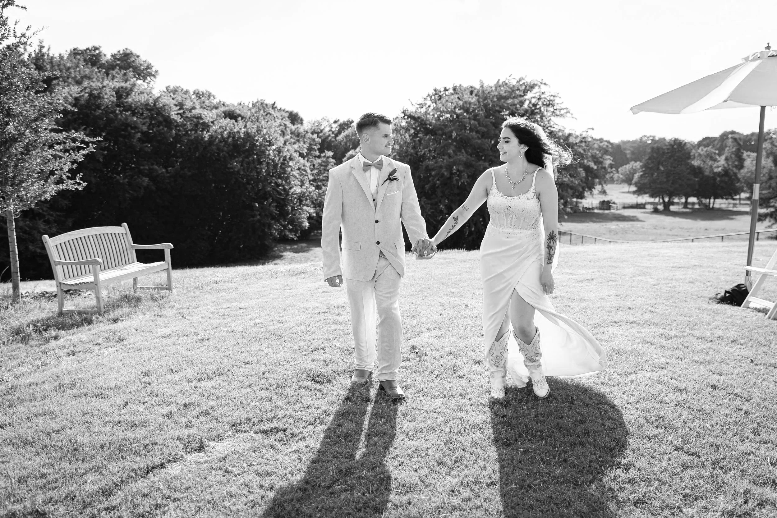 A black and white photo of a couple holding hands walking across a grassy field outdoors. The man is wearing a suit and bow tie, and the woman is in a wedding dress with tattoos on her arms. There are trees and a bench in the background and an umbrella to the right.