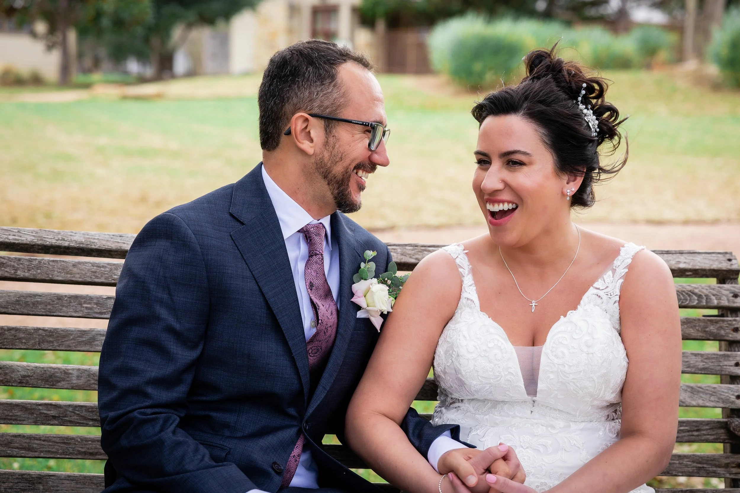 A bride and groom sitting on a park bench, smiling and enjoying each other's company outdoors.