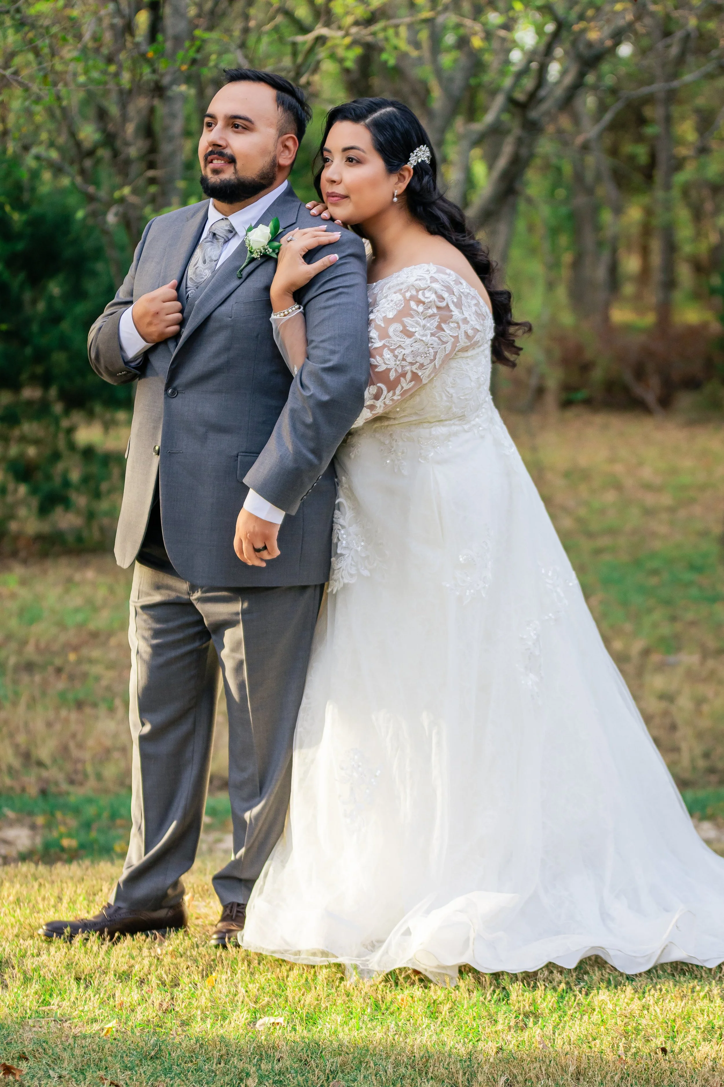 A bride and groom standing outdoors on grass, with trees in the background, during a wedding photoshoot. The groom is in a gray suit with a white shirt, patterned tie, and boutonniere, and has a beard and short dark hair. The bride is wearing a white lace wedding dress with long lace sleeves, pearl earrings, a hair accessory, and has dark wavy hair.
