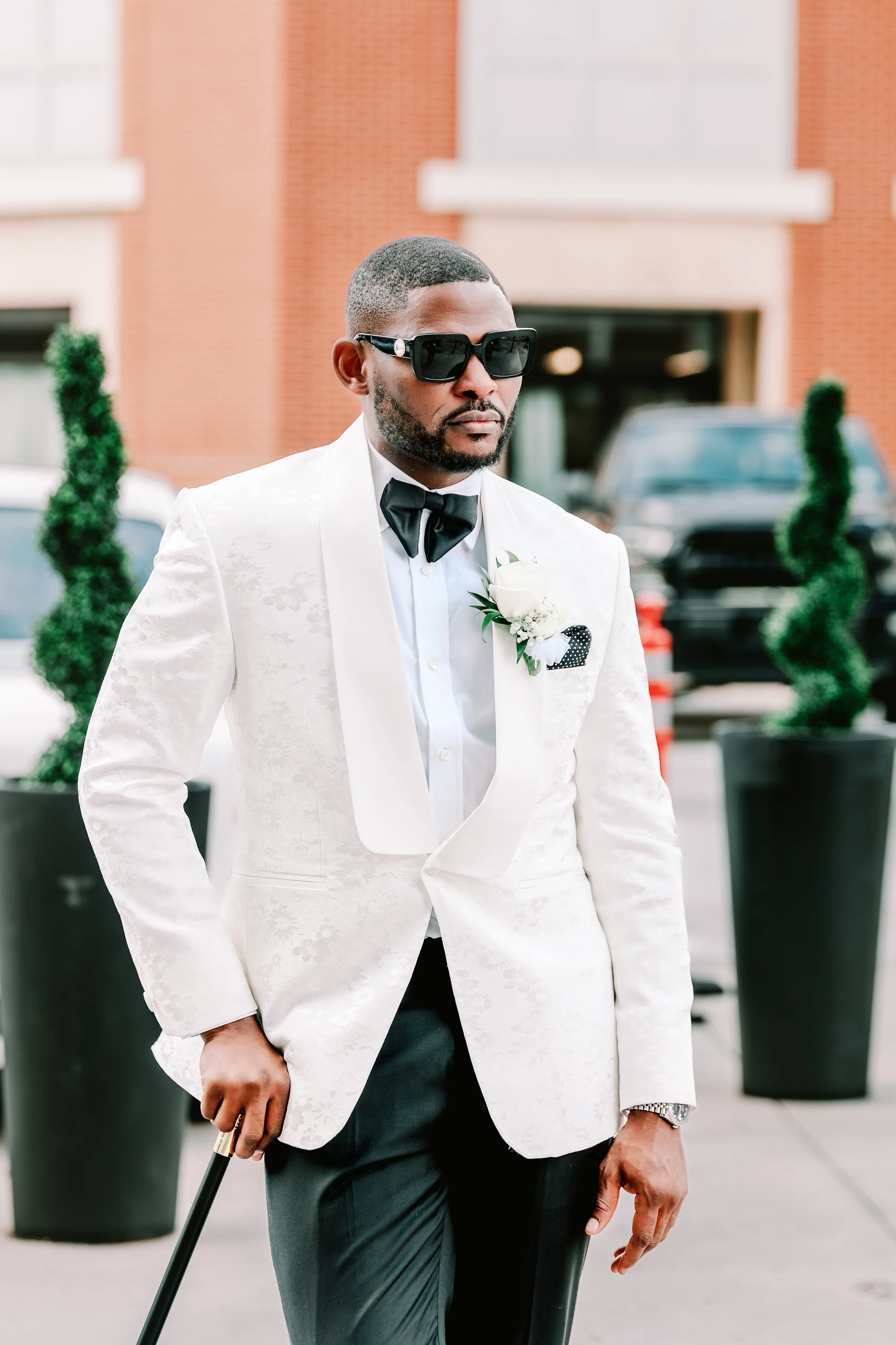 A man wearing a white tuxedo jacket with floral pattern, black dress pants, a white shirt, and a black bow tie, standing outdoors. He has a boutonniere with white flowers and greenery on his left lapel, black sunglasses, and a wristwatch. He holds a cane in his right hand and has neatly groomed facial hair. The background shows potted topiary plants and a brick building.