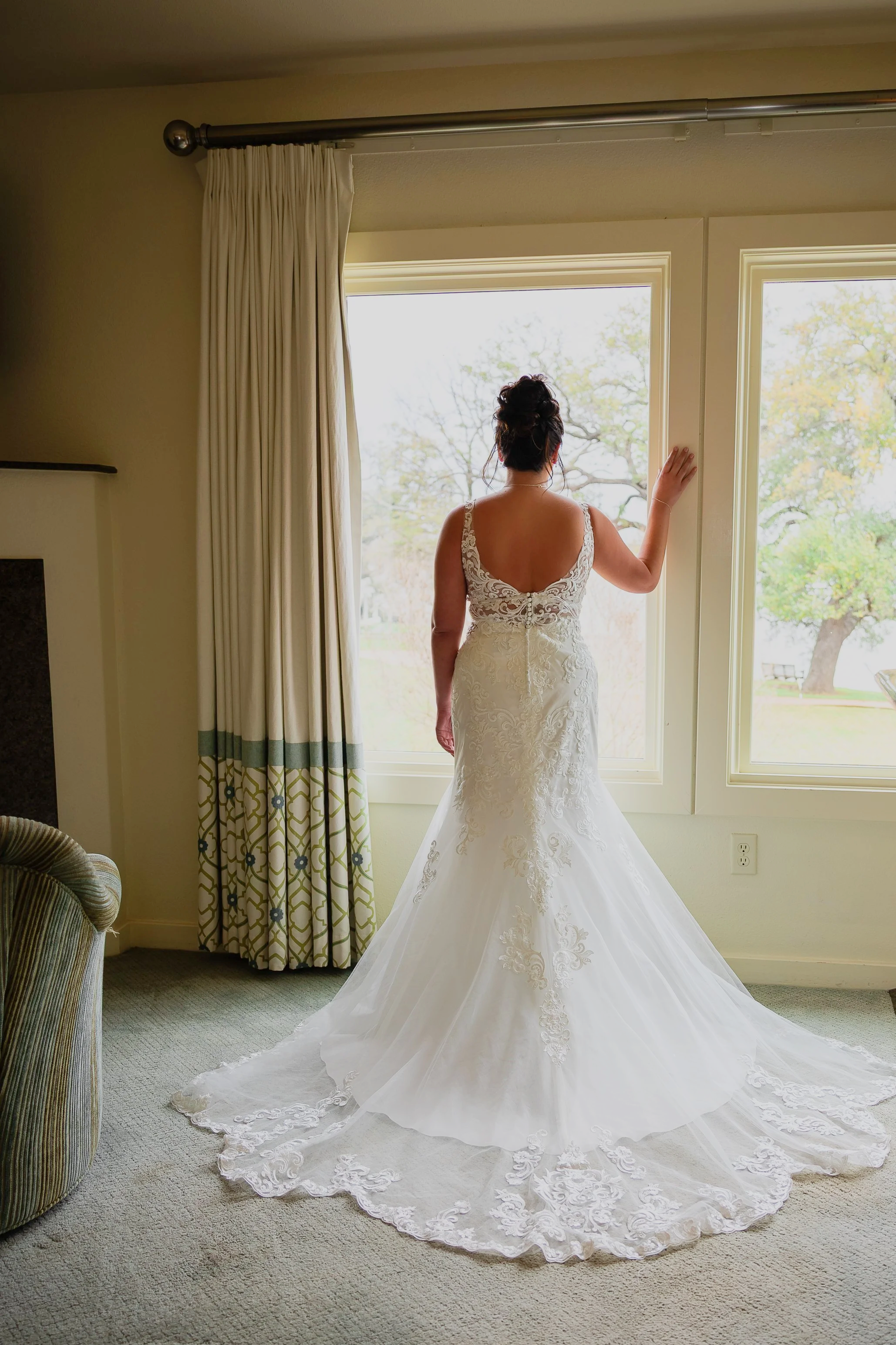 A woman in a wedding dress stands in front of large windows, facing away, with her hand resting on the window frame. The dress has lace details on the back and a long train on a light-colored carpeted room.