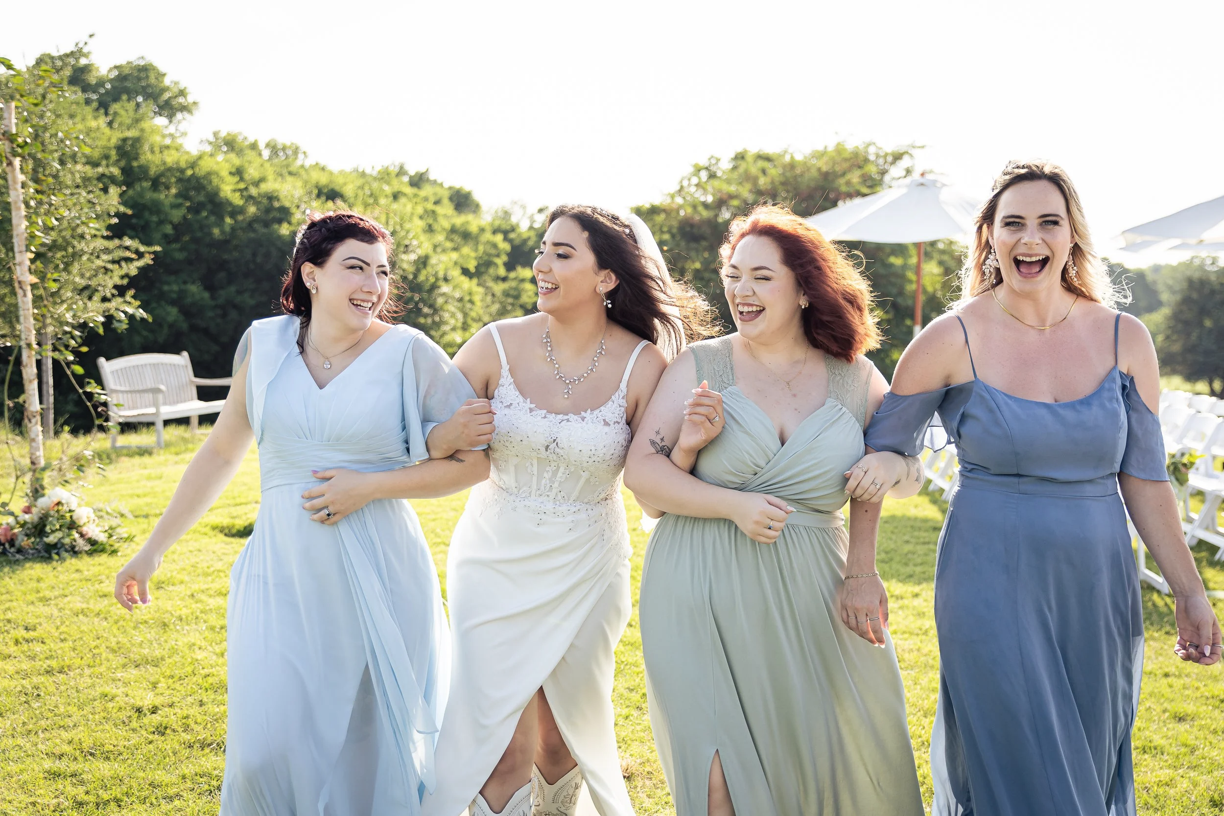 Four women in formal dresses happily walking arm in arm outdoors on a sunny day, with a green background and white umbrellas in the distance.