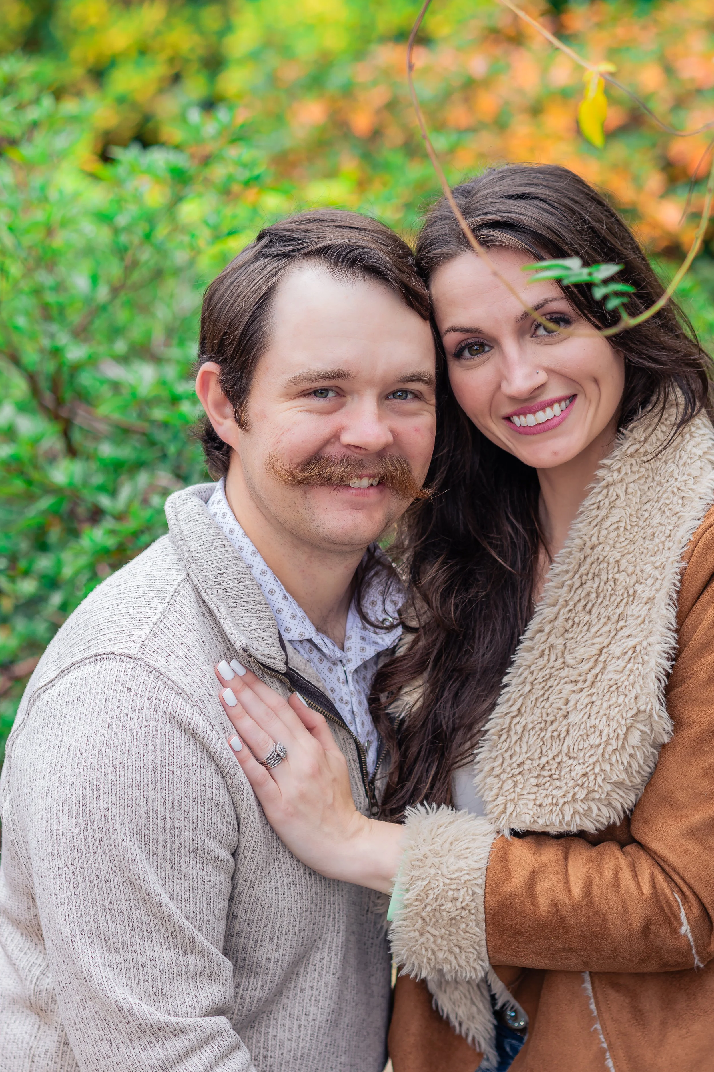 Close-up of a smiling couple embracing outdoors surrounded by fall foliage. The man has a mustache, light hair, and a light-colored sweater. The woman has long dark hair, a nose piercing, and a cream furry jacket.
