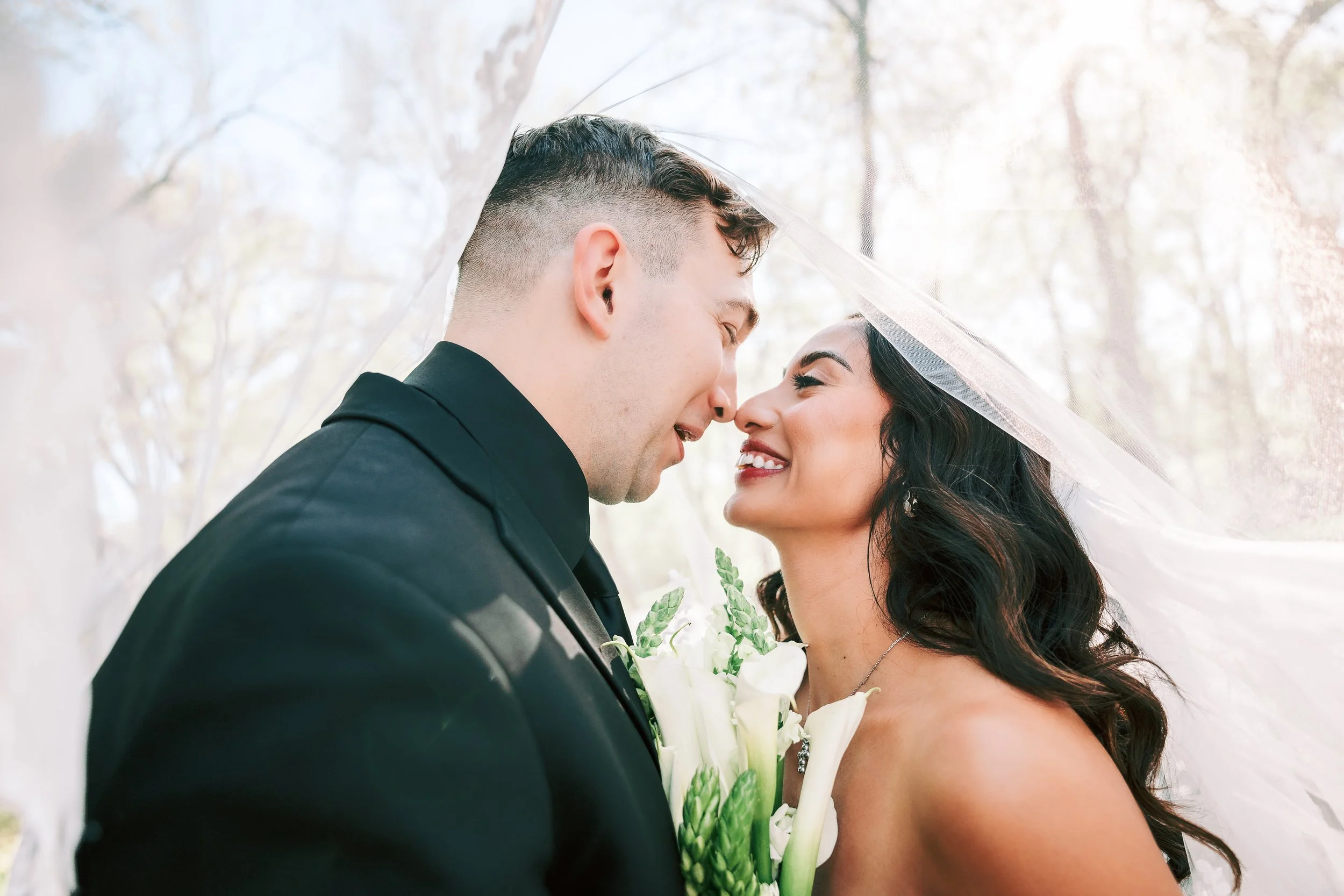A bride and groom on their wedding day, smiling and leaning into each other under a translucent umbrella in an outdoor setting with blurred trees in the background.