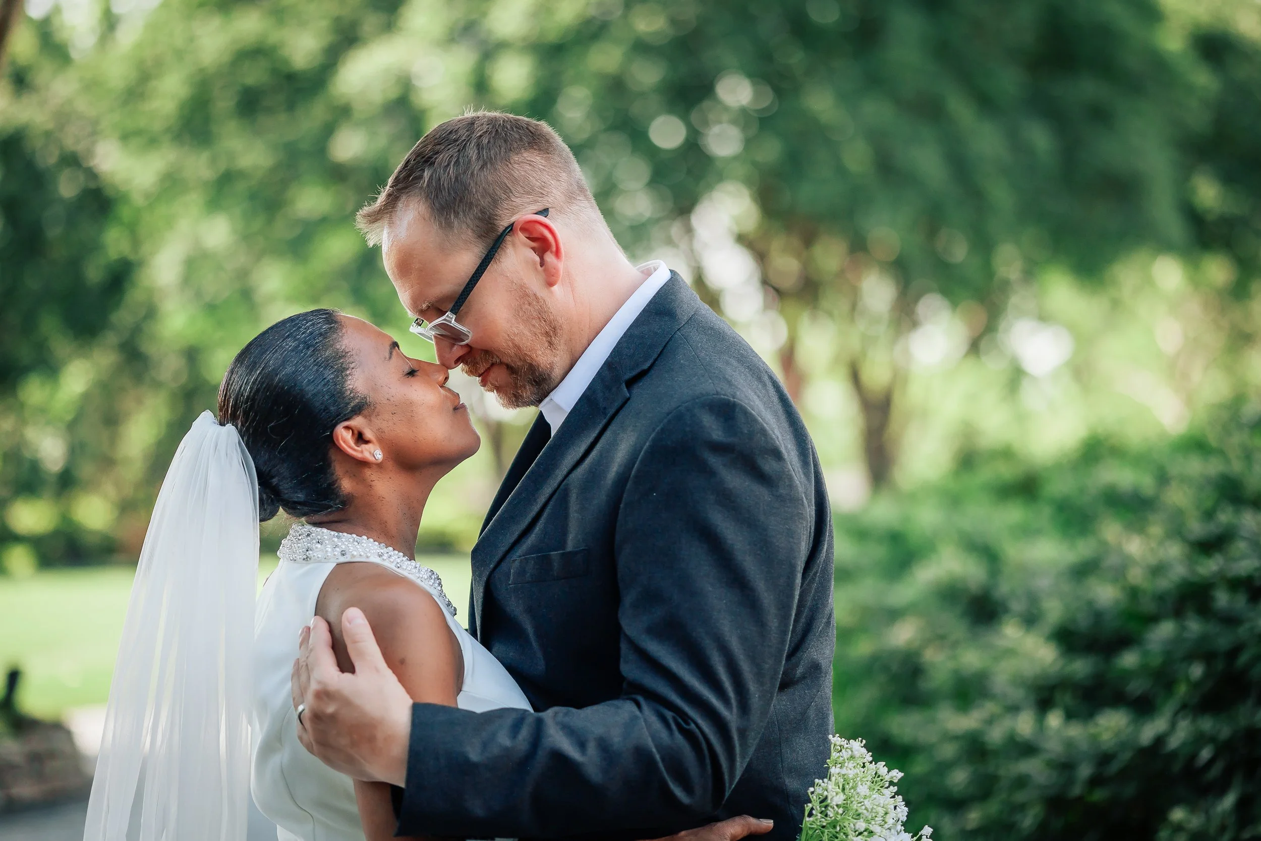 A bride and groom share an intimate moment outdoors, touching foreheads with eyes closed, in a green park setting.