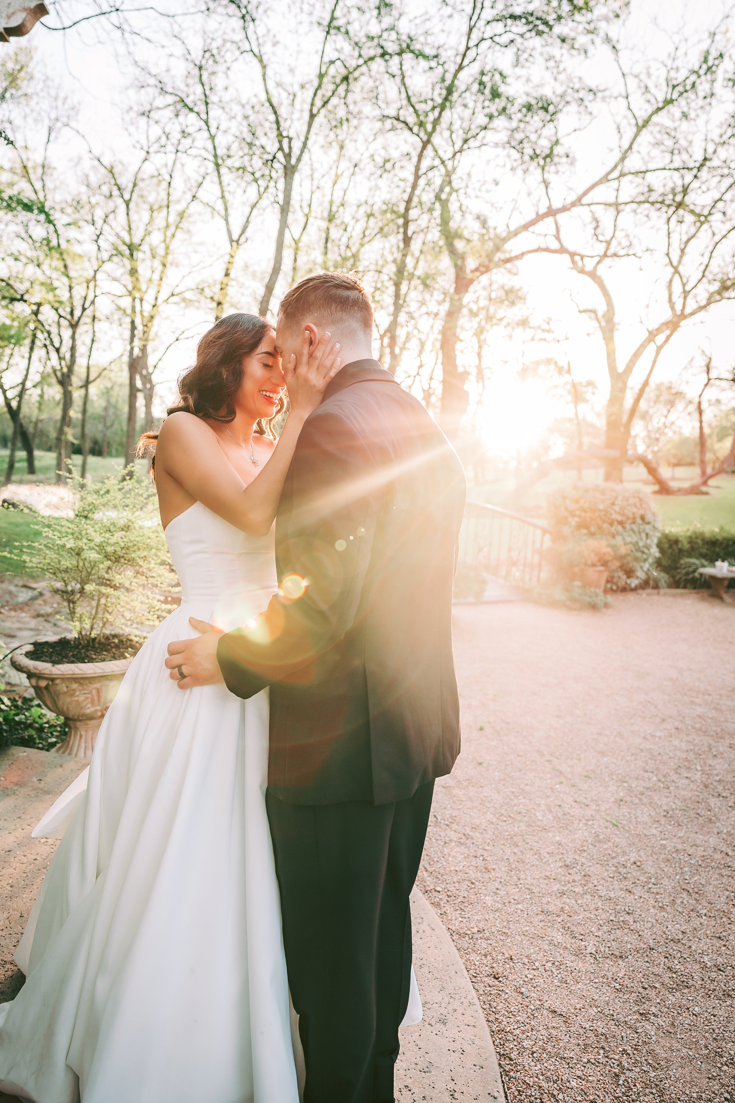 A couple dressed in wedding attire embracing outdoors during sunset, with trees in the background.