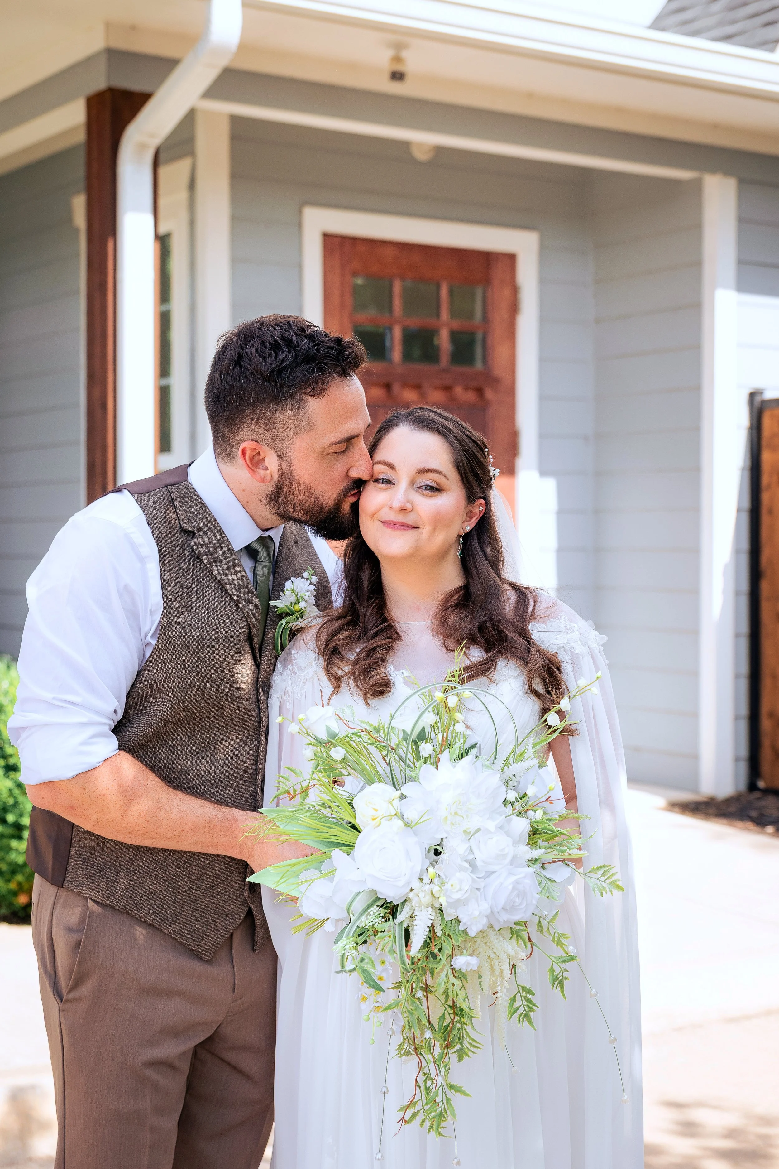 A groom kisses a bride on her cheek at their wedding outside a house, with the bride holding a large bouquet of white flowers and greenery.