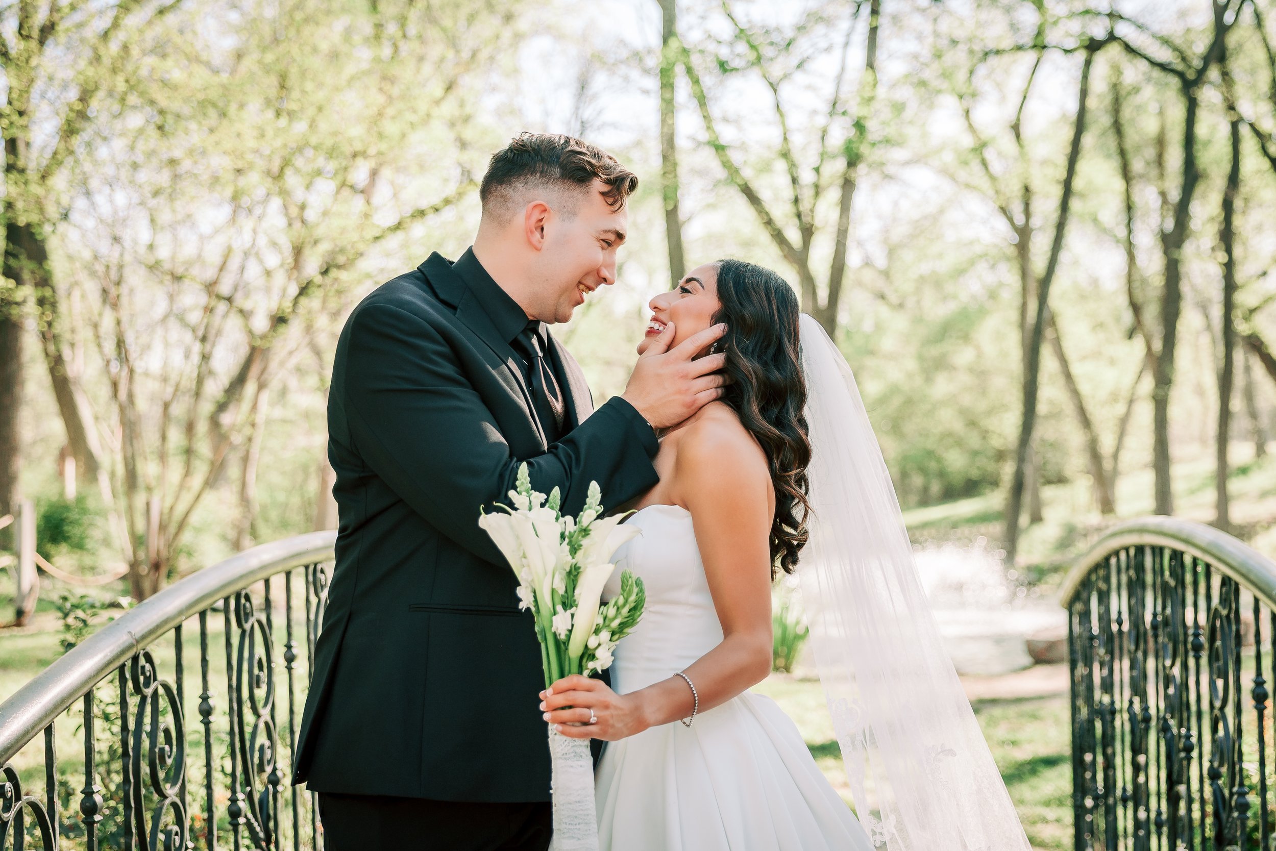 A happy bride and groom standing on a bridge in a park, smiling at each other during a wedding photo shoot. The groom is wearing a black suit, and the bride is dressed in a wedding gown holding a bouquet of white calla lilies. The background features trees with green leaves and sunlight filtering through.