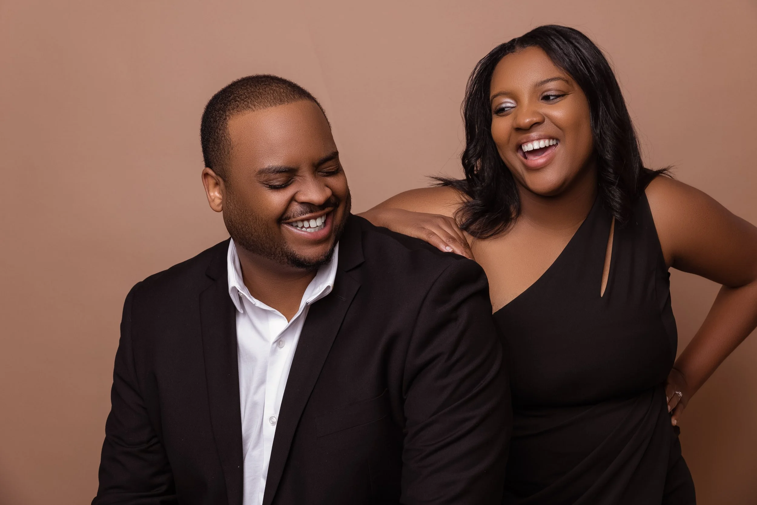 Two people, a man and a woman, are smiling and laughing together against a plain beige background. The man is wearing a dark suit with a white shirt, and the woman is wearing a black sleeveless top. They appear to be enjoying a happy moment.