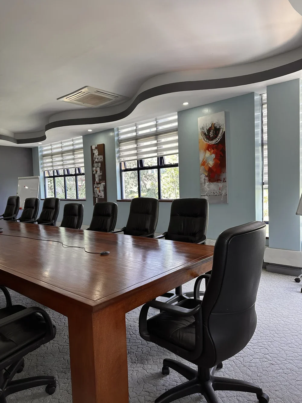 Empty conference room with a long wooden table surrounded by black leather office chairs, windows with horizontal blinds, and modern artwork on the light blue walls.