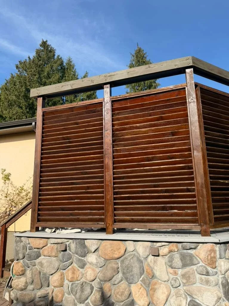 Wooden balcony with horizontal slats on stone foundation, with trees and a blue sky in the background.