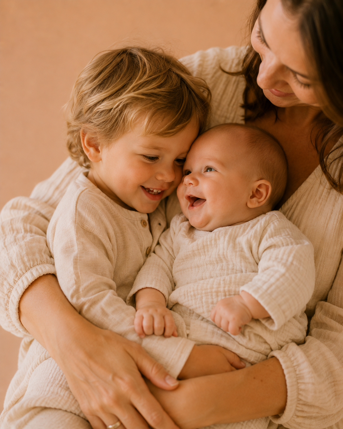 A woman holding two young children, a boy and a baby, all smiling happily and close together.