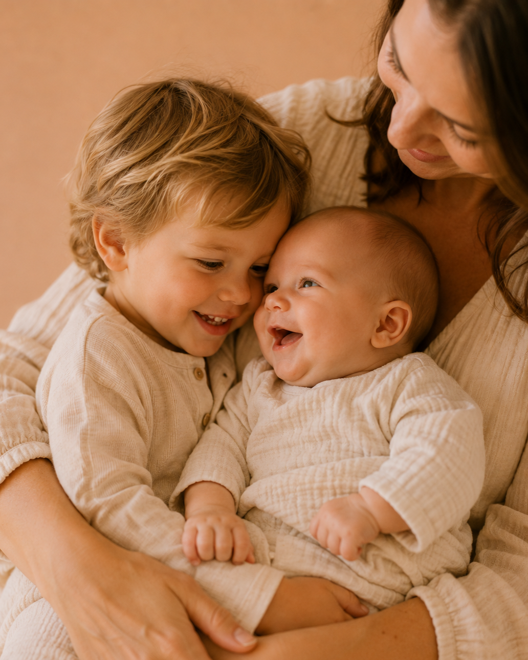 A woman holding two young children, a boy and a baby, all smiling happily and close together.