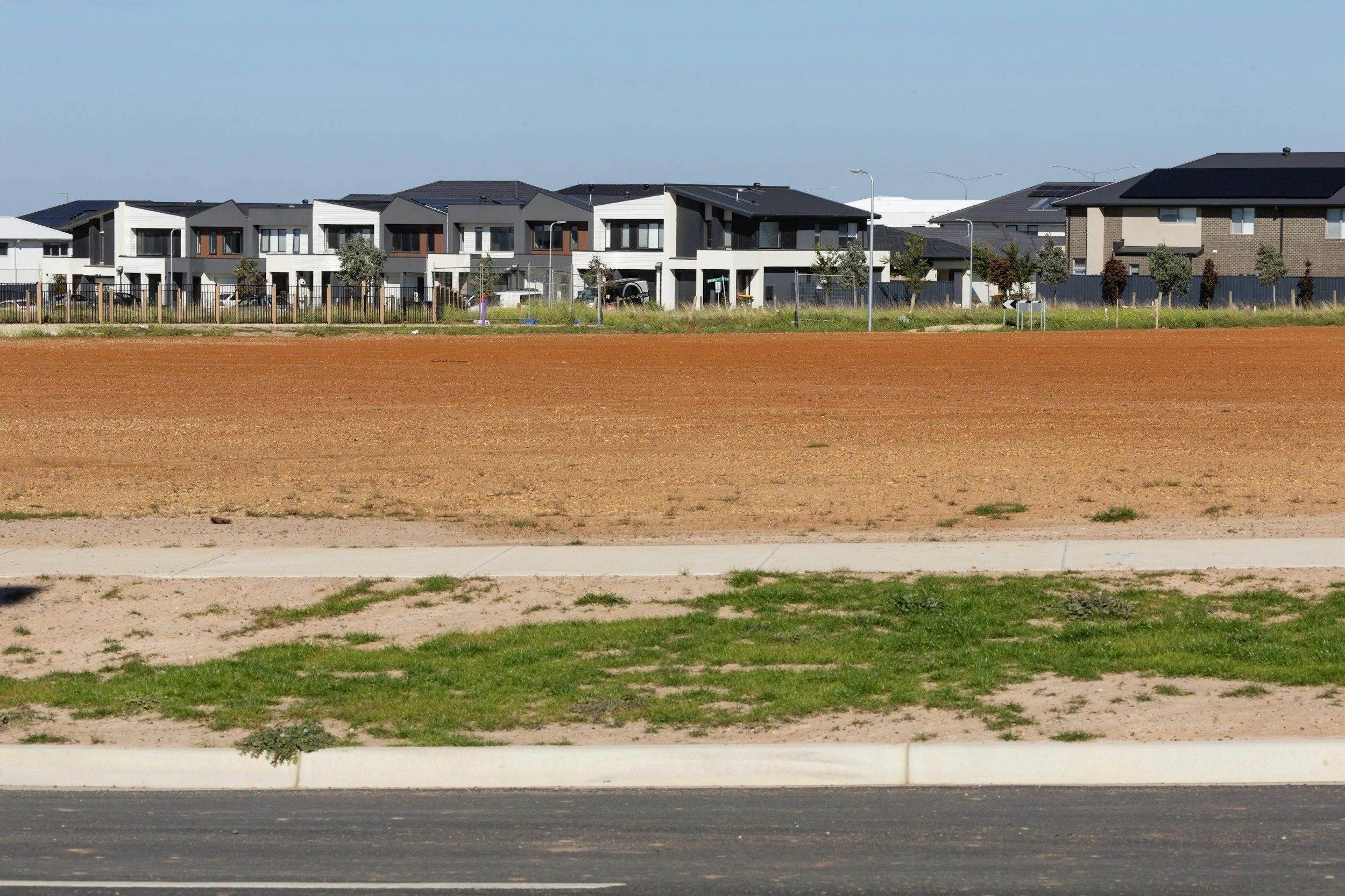 A street view with a foreground of asphalt road, middle section of grass and dirt, and background of modern residential houses with dark roofs and some with solar panels, under a clear blue sky.