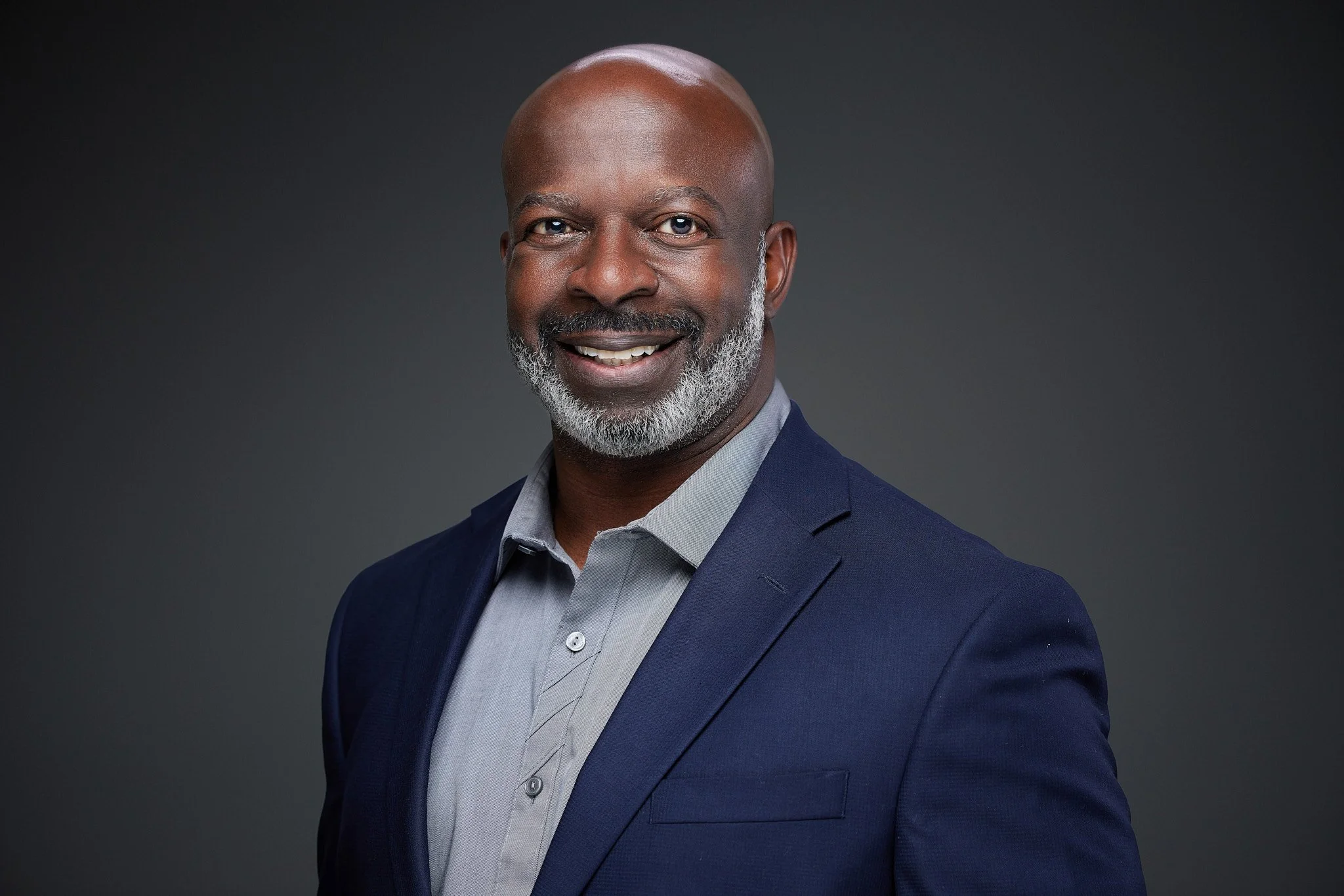 A portrait of a bald, middle-aged African American man with gray beard, wearing a navy blazer over a light shade dress shirt, smiling against a dark gray background.