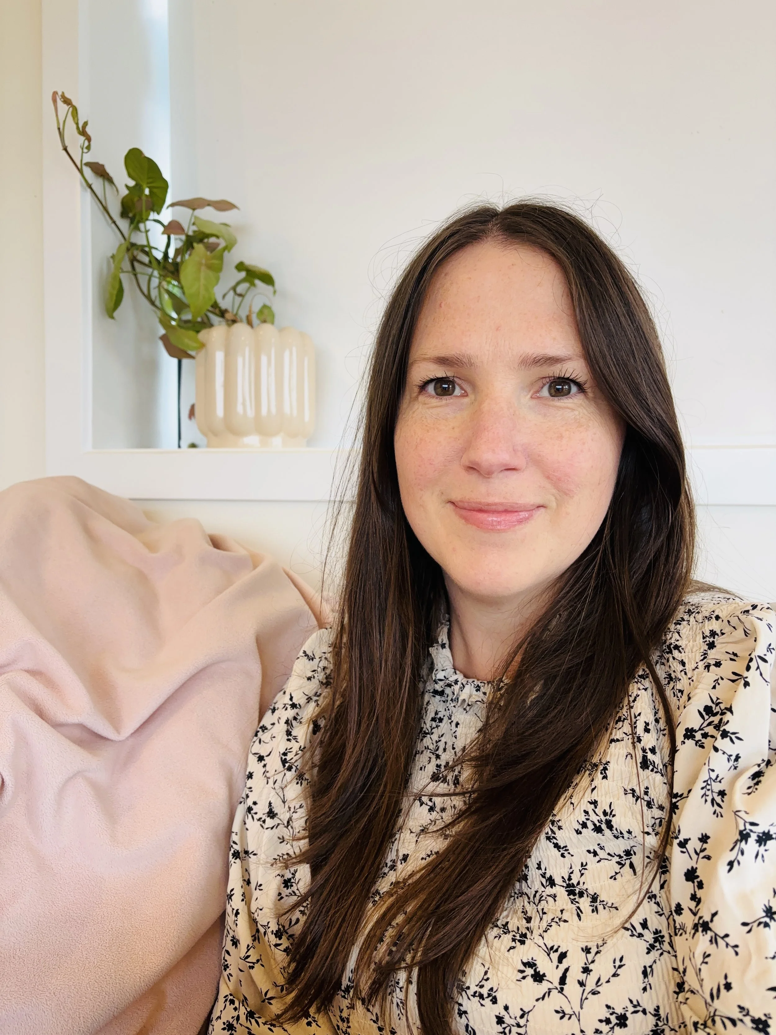 A woman with long brown hair and freckles smiling at the camera, wearing a floral dress. She is sitting next to someone wearing a light pink sweater, in front of a white wall with a small window that has a potted plant on the windowsill.