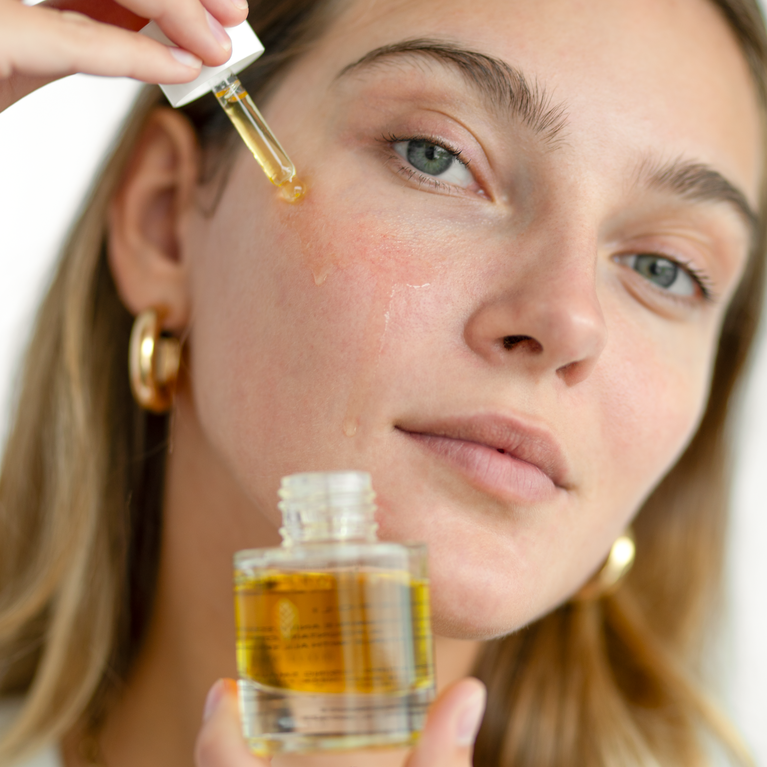 A woman with blonde hair and green eyes holding a small glass bottle with a dropper, dispensing a yellow liquid onto her face as part of a skincare routine. Toronto luxury botanical facials. Toronto organic facials.