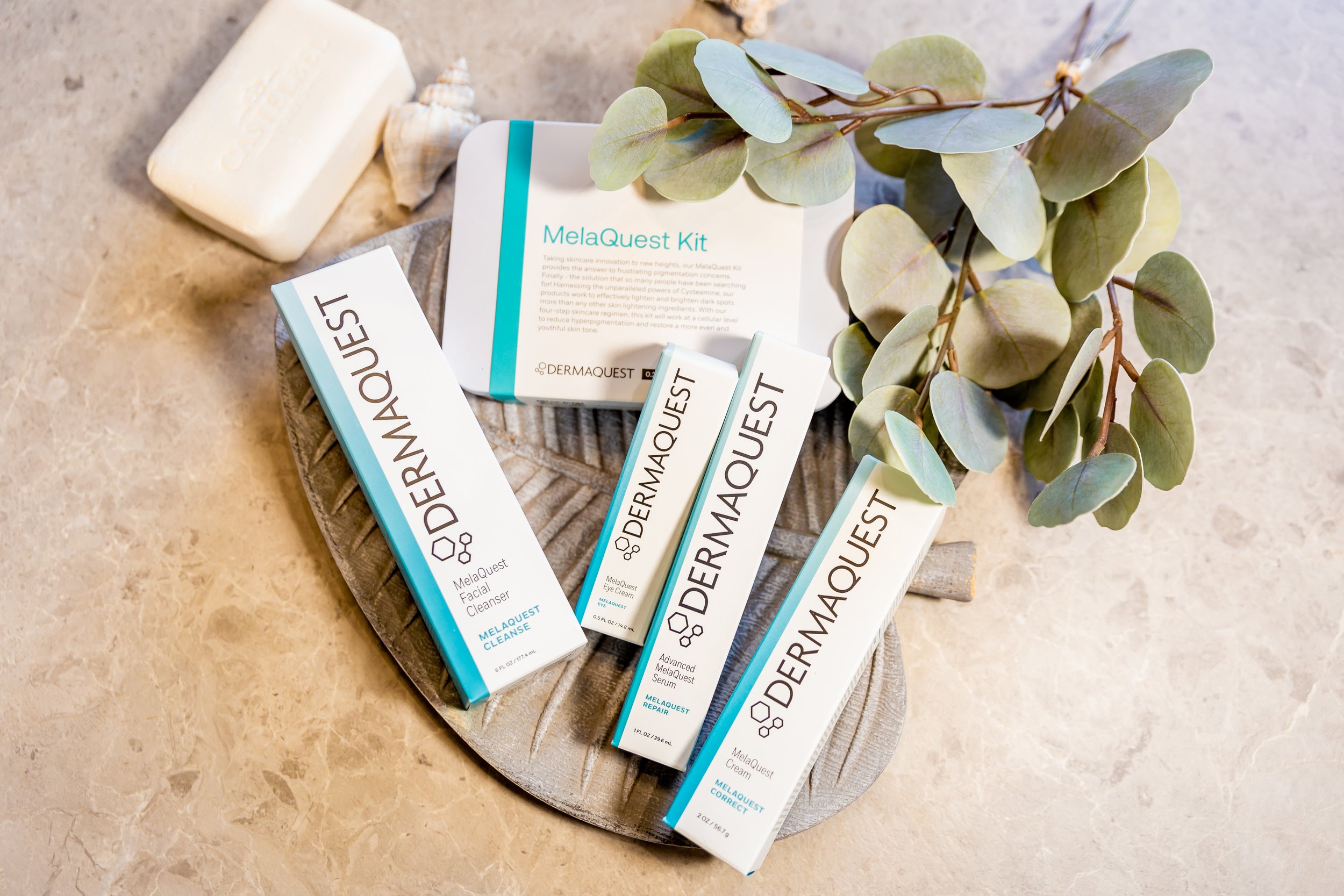 A flat lay of Dermaquest skincare products, including facial cleanser, eye cream, and serum, arranged on a decorative plate with green eucalyptus leaves and a bar of soap nearby, all on a beige marble surface. Toronto medical grade skincare.
