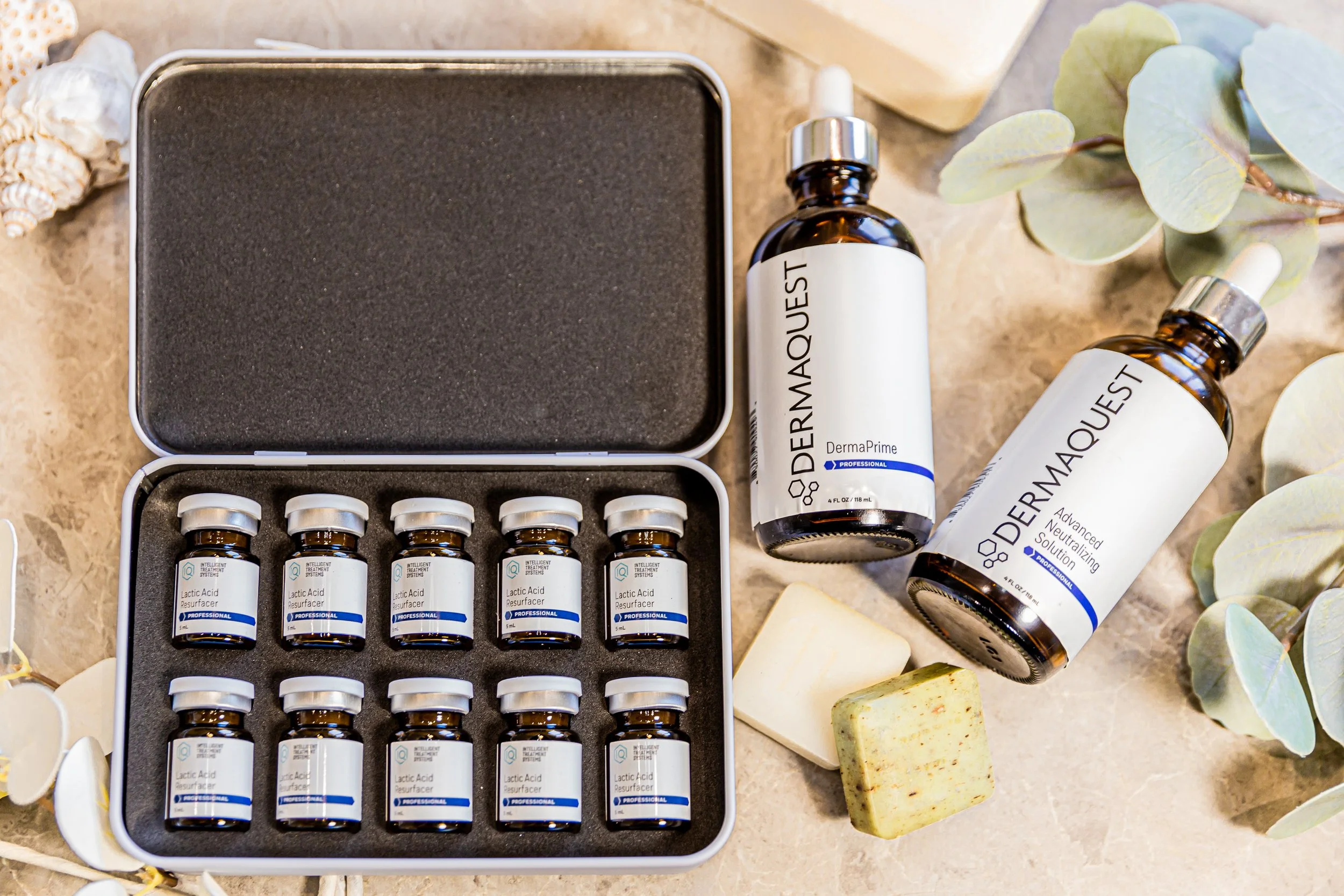 Open box of small amber glass bottles labeled 'Lactic Acid Resurfacer' arranged in rows. There are two more bottles outside the box labeled 'DermaQuest Advanced Neutralizing Solution'. Additional items include soap bars, seashells, and green leaves for decoration.