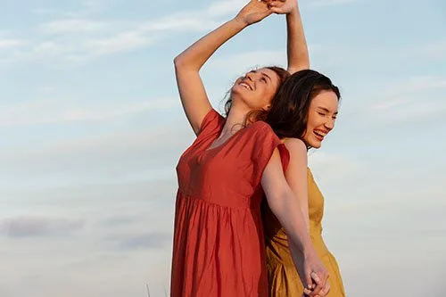 2 jeunes femmes souriantes, lâcher prise, dos à dos, bras vers le ciel qui célèbrent la vie