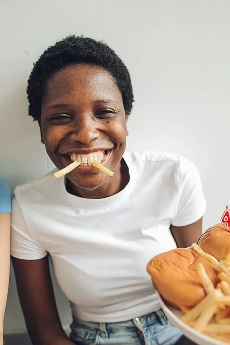 Une femme souriante avec des frites dans la bouche et un plateau de nourriture avec frites et burgers