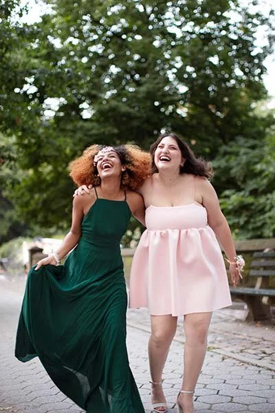 Deux jeunes femmes qui rient et marchent ensemble dans un parc, l'une en robe verte et l'autre en robe rose, entourées d'arbres et de nature.