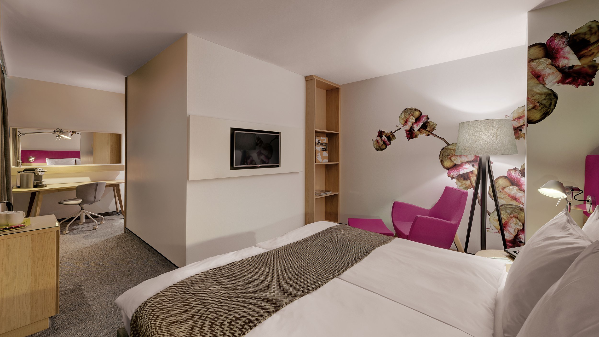 A hotel room with a bed, pink armchair, floor lamp, and a wall with floral wallpaper, with a workspace area visible in the background.
