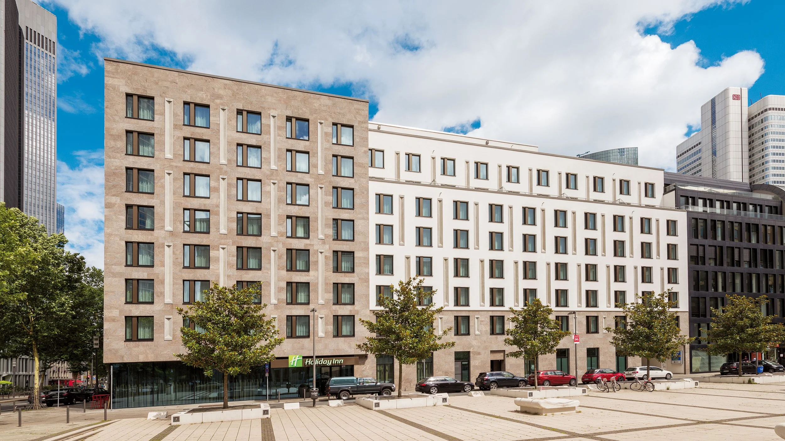 Modern multi-story hotel building with storefront entrance, parked cars, bicycles, trees, and a partly cloudy sky in an urban setting.