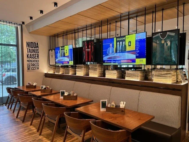 Interior of a cafe with wooden tables and chairs, a long booth with a cushioned back, a row of newspapers or magazines above the booth, four TV screens displaying news and sports, large window on the left, and a wall with text listing cities.